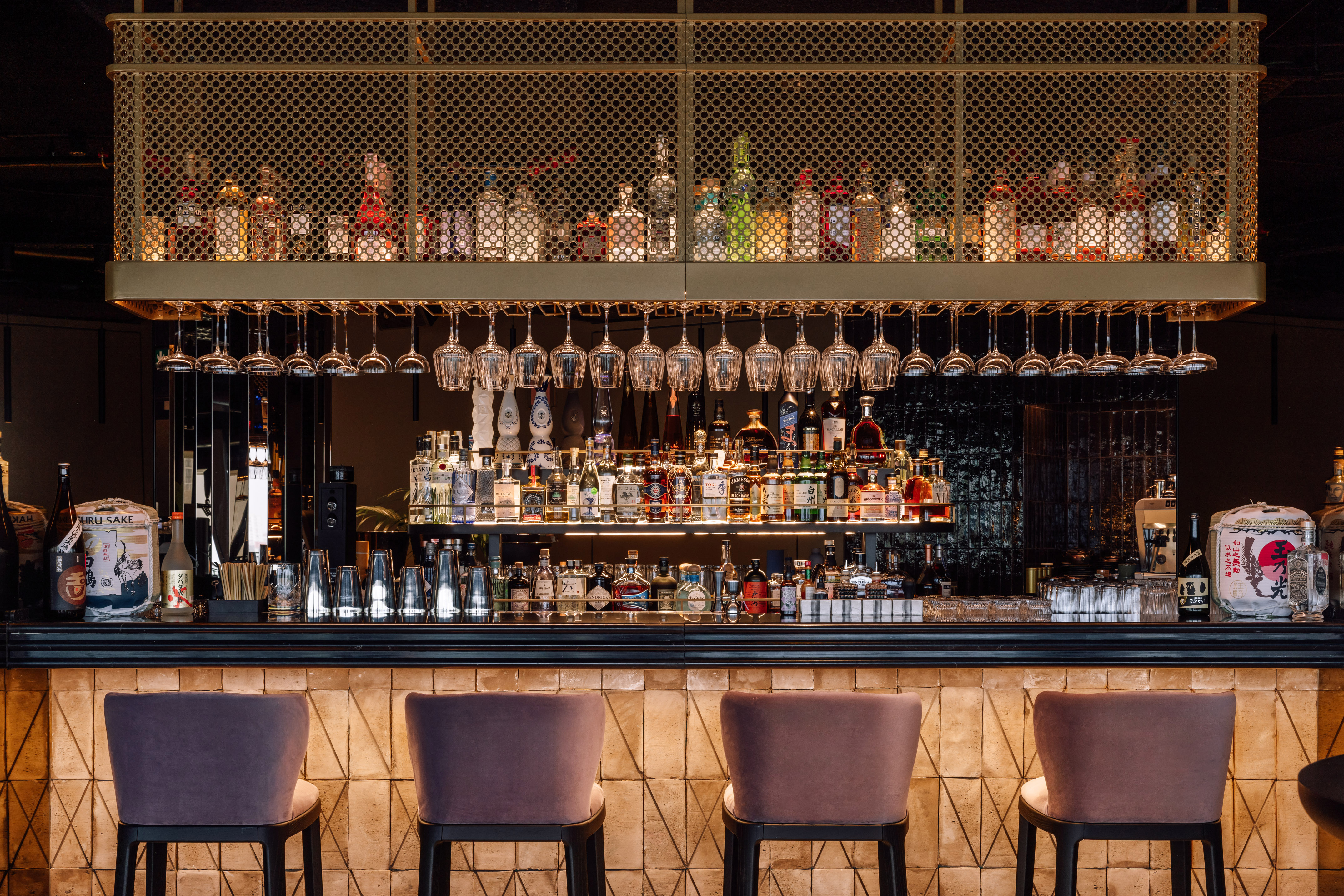Modern bar with diverse liquor, hanging glasses, and velvet stools.