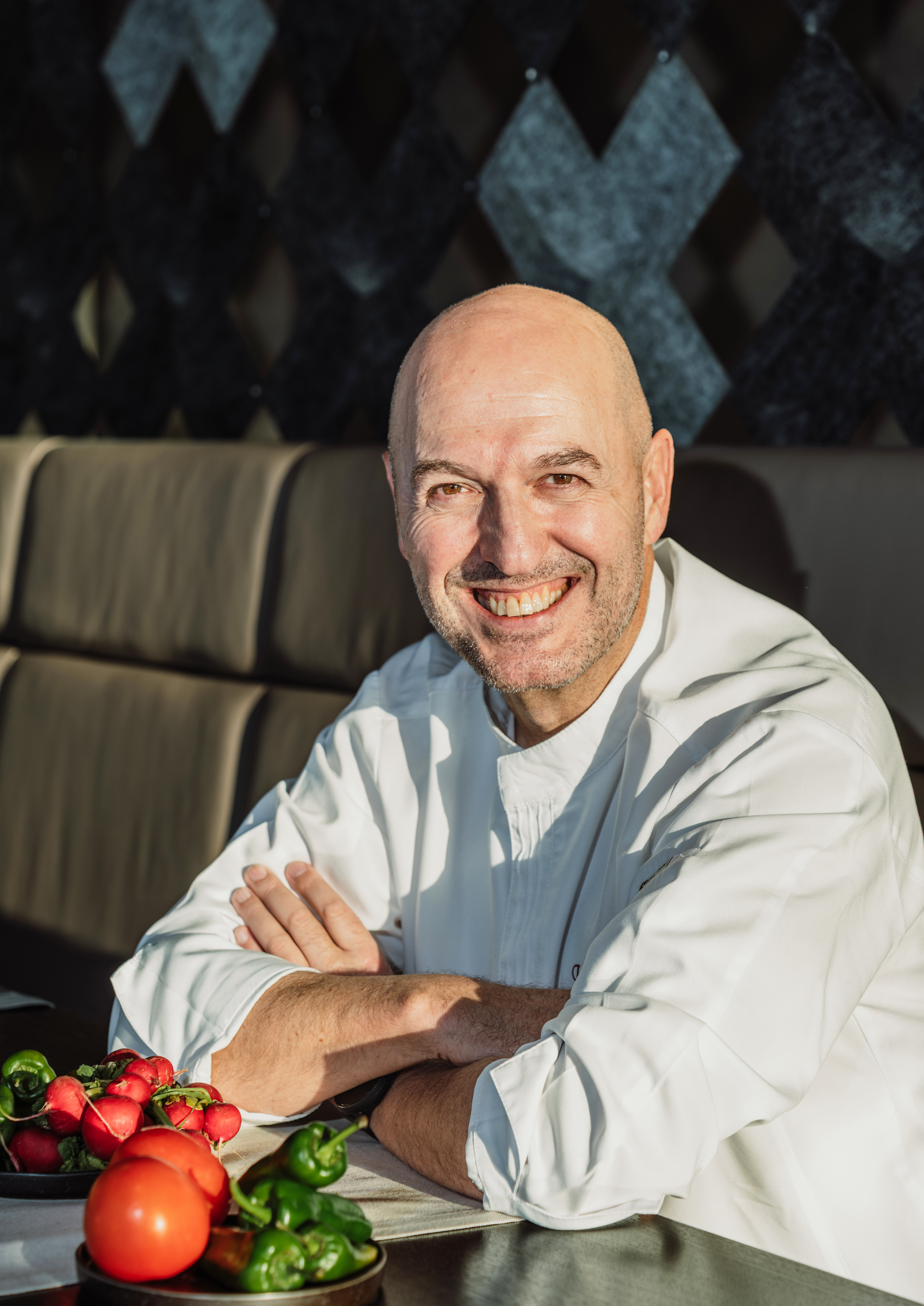 Bald chef smiling in white uniform, fresh produce on table.