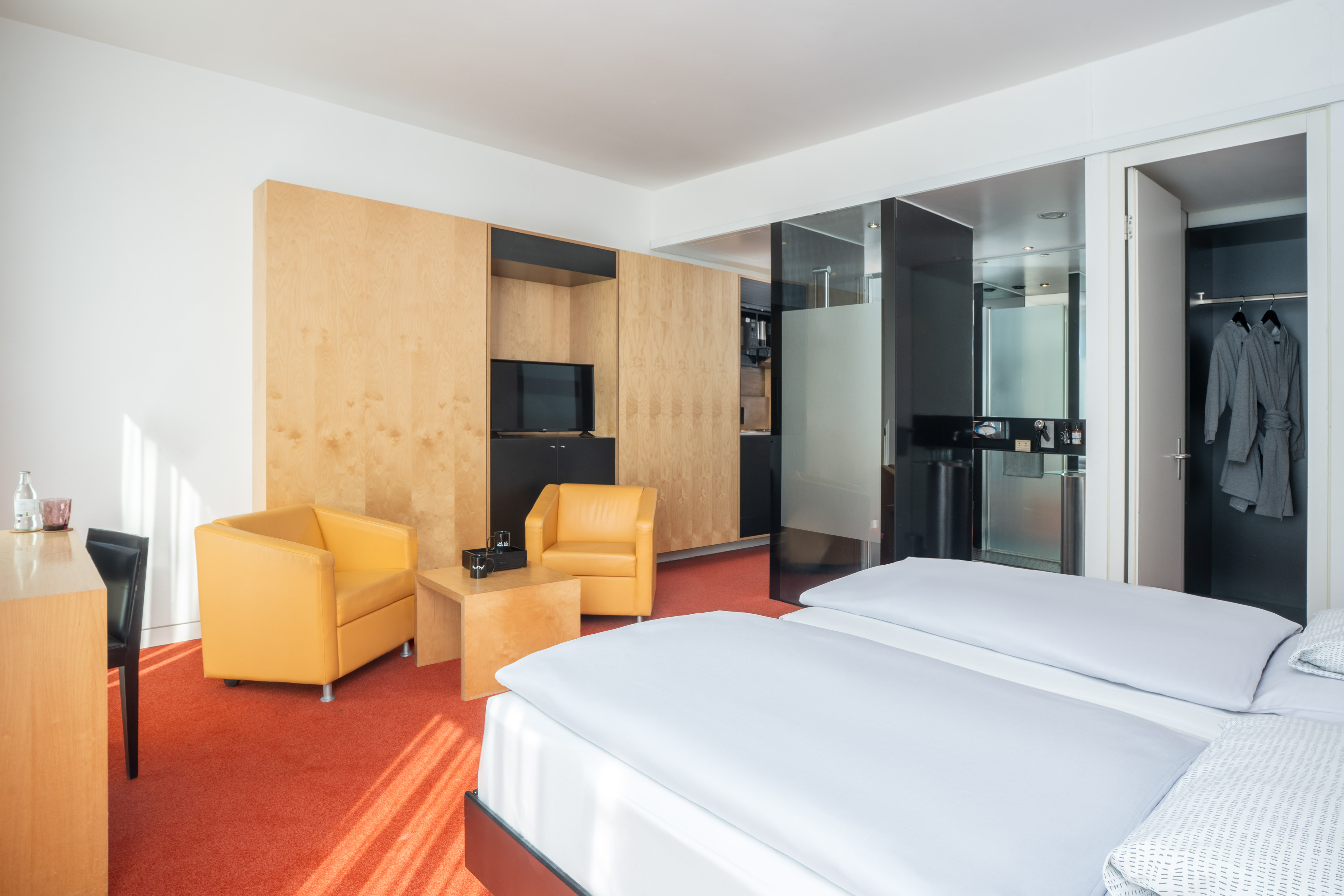 Modern hotel room with twin beds, yellow armchairs, wood storage, and red carpet.