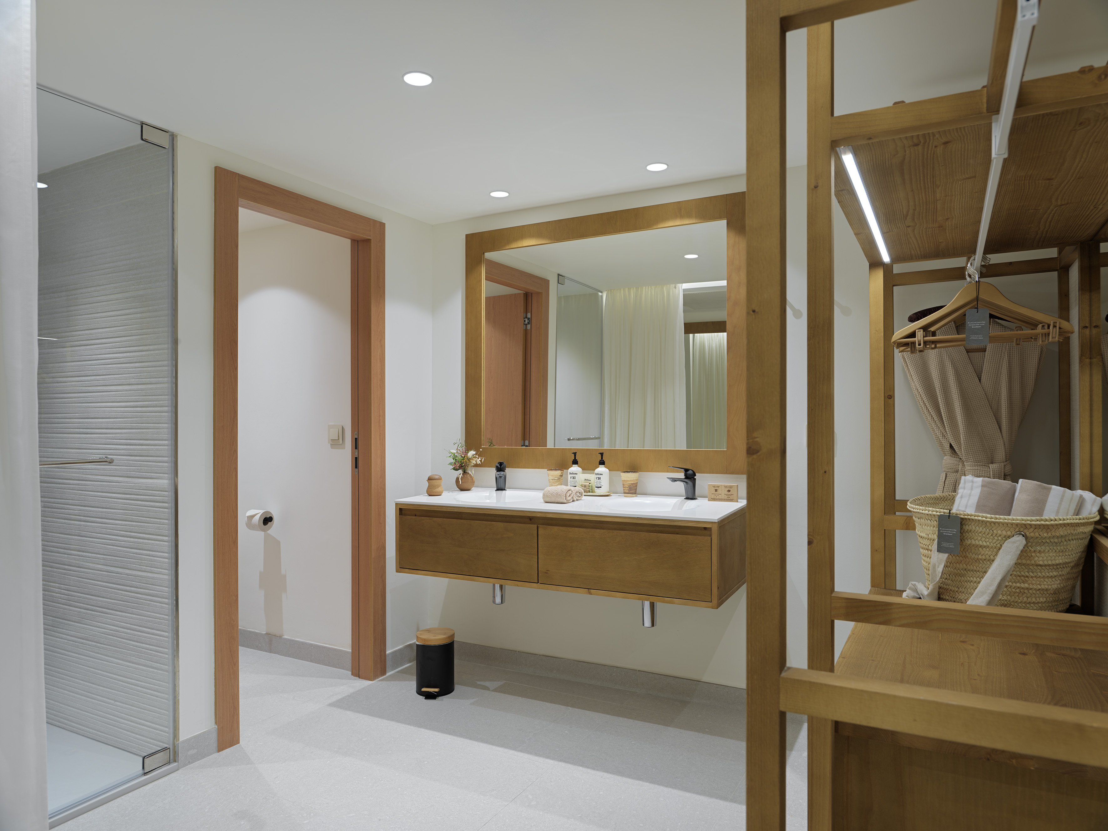 Modern hotel bathroom with wooden double vanity, glass shower, and open closet.