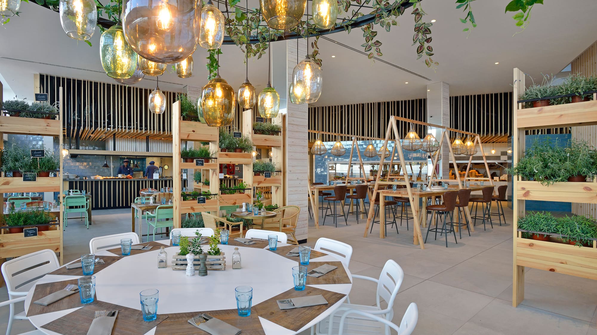 Modern restaurant with lush greenery, warm hanging lights, and open kitchen.
