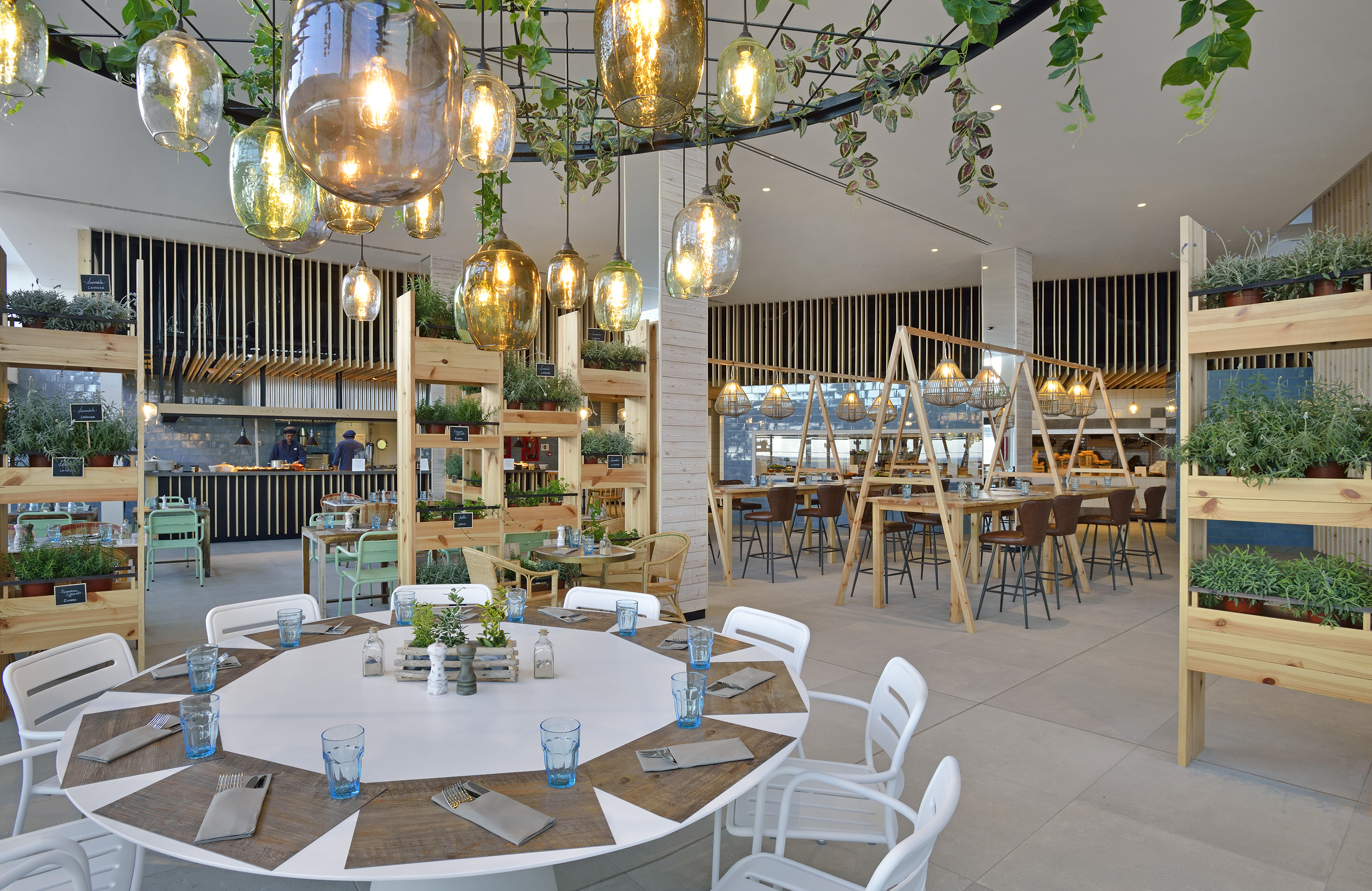 Modern restaurant with lush greenery, warm hanging lights, and open kitchen.