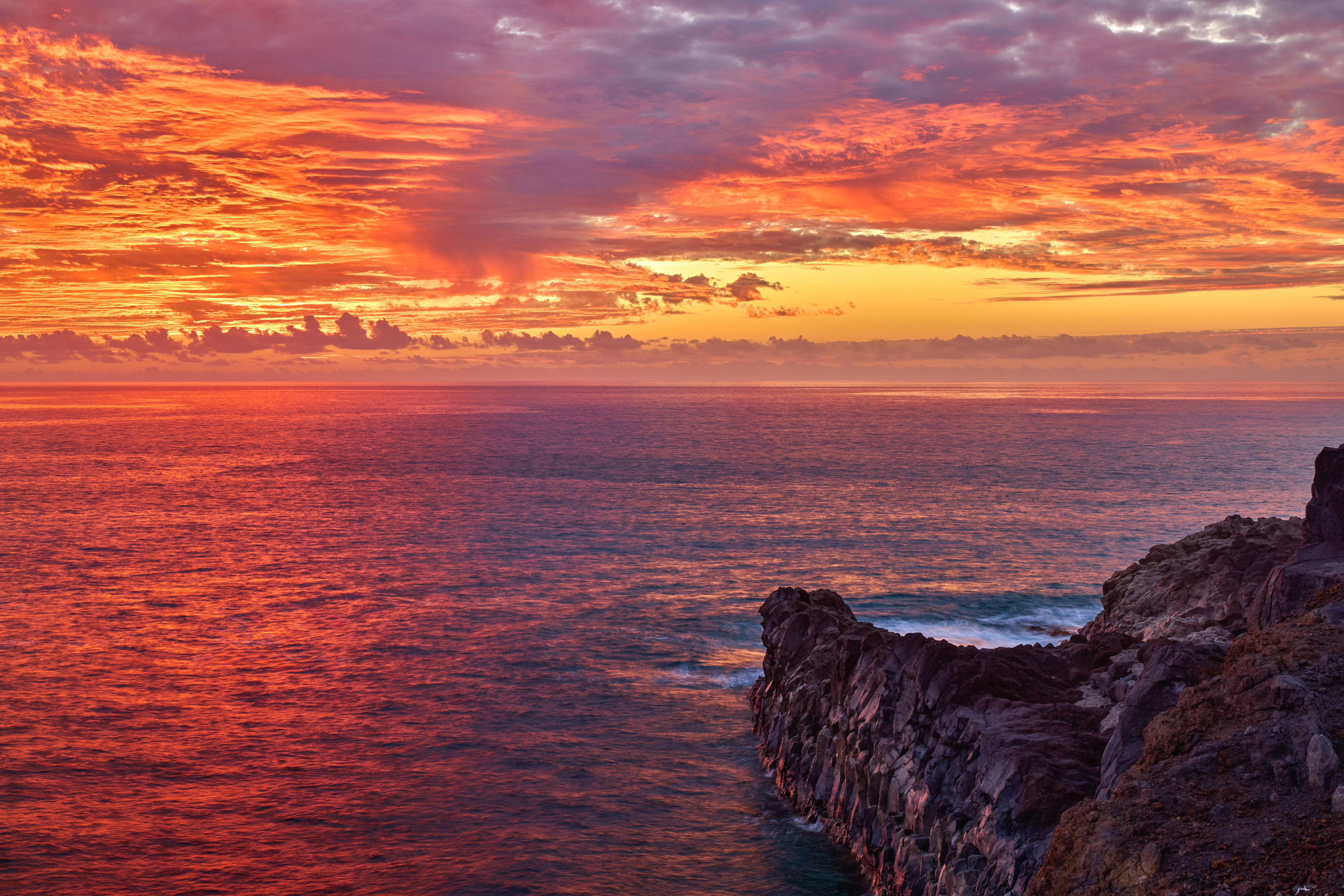 Fiery sunset over a red ocean, dark rocky cliff along the coast.