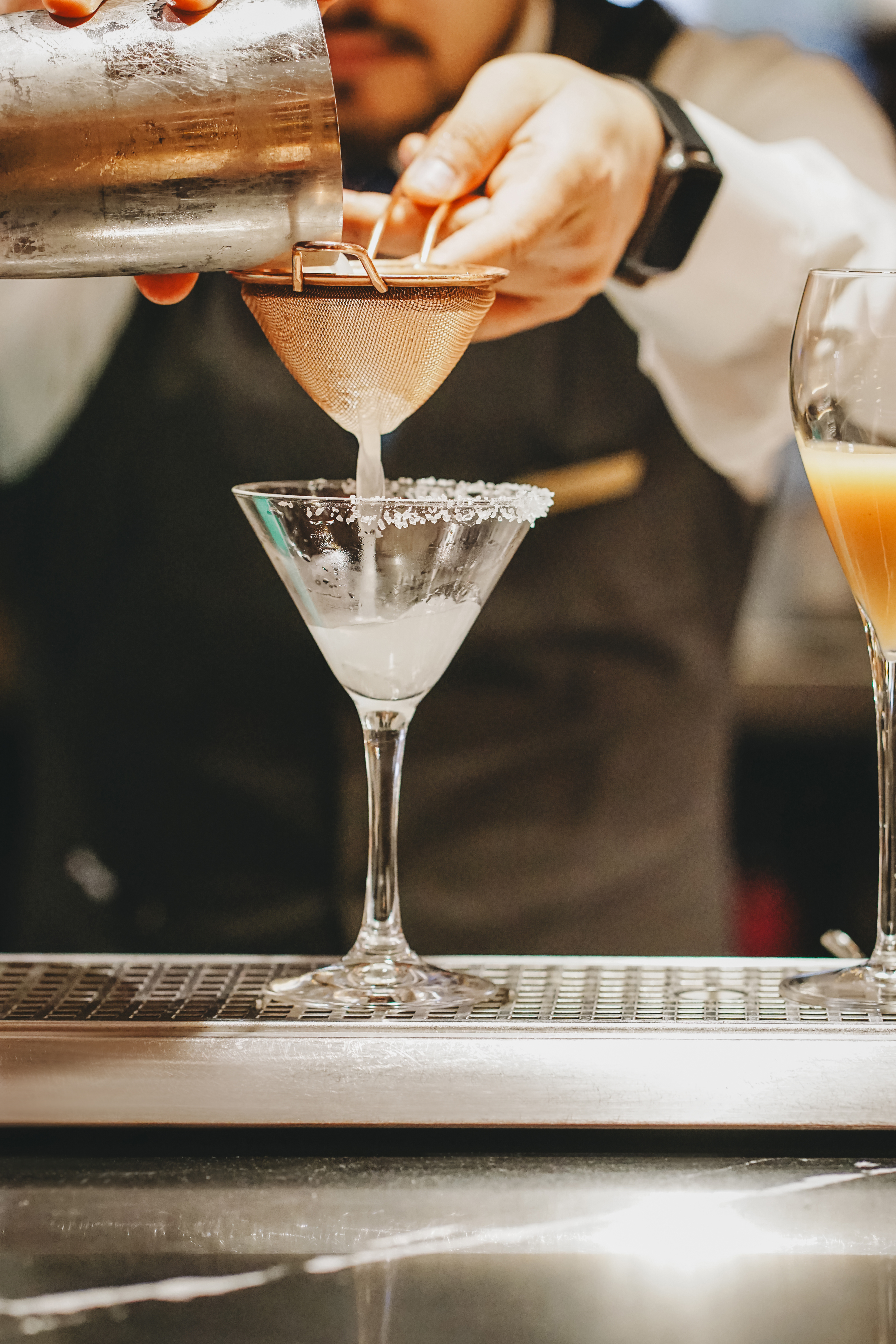 Bartender straining cocktail into salt-rimmed martini glass.