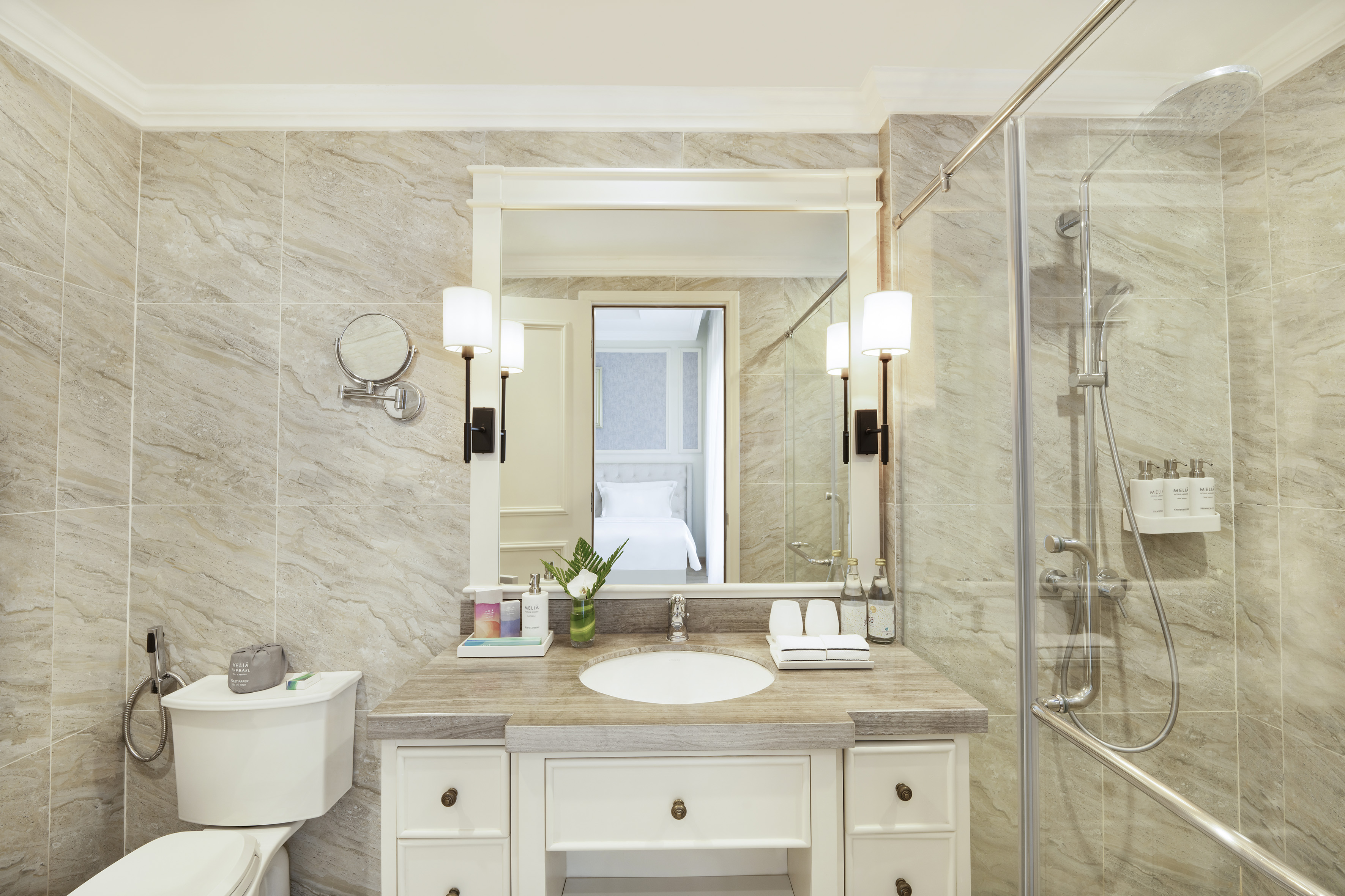 Bright hotel bathroom with vanity, mirror, shower, toilet, and marble-look tiles.