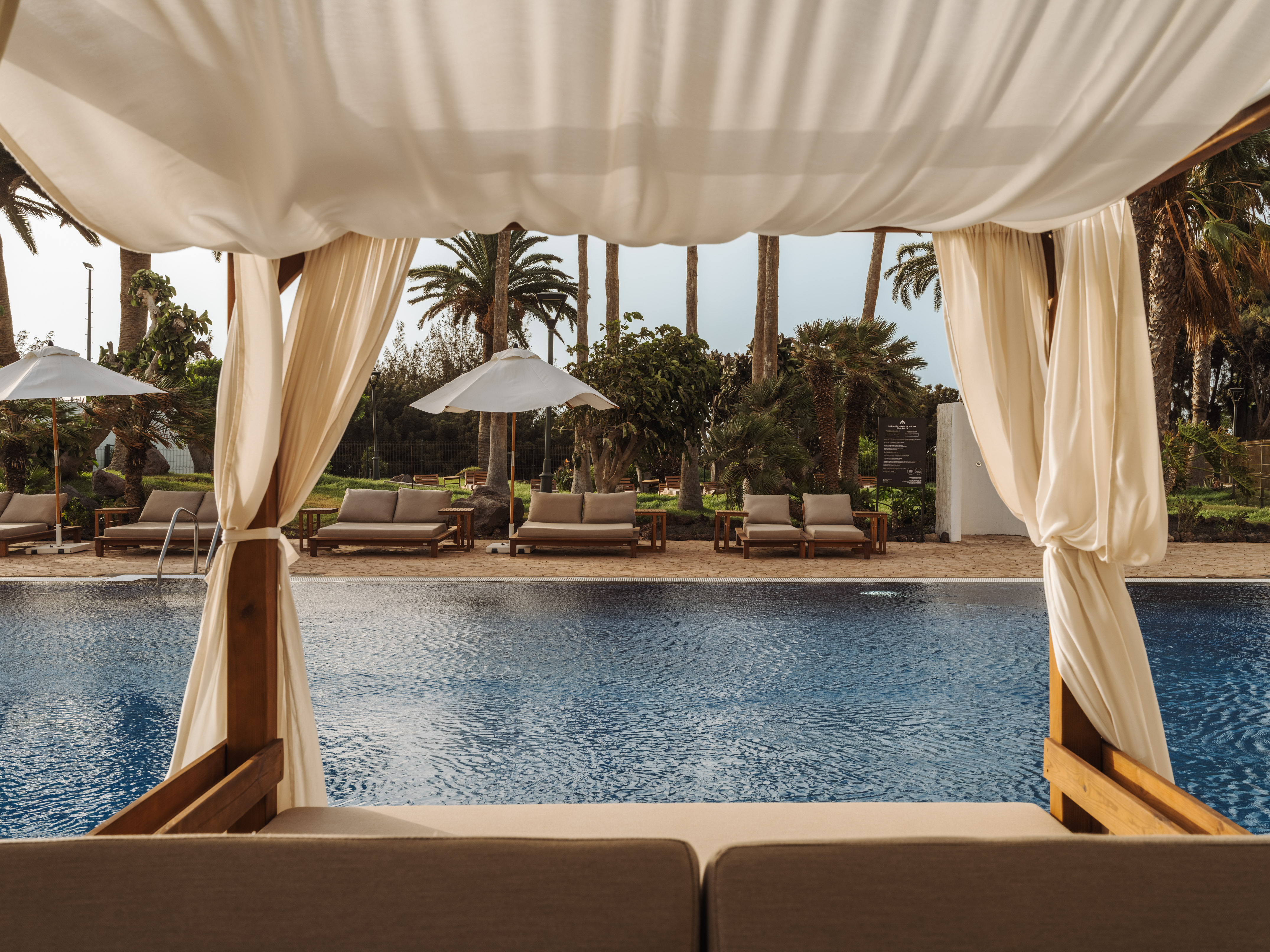 Resort swimming pool view from white cabana, palm trees, lounge daybeds.