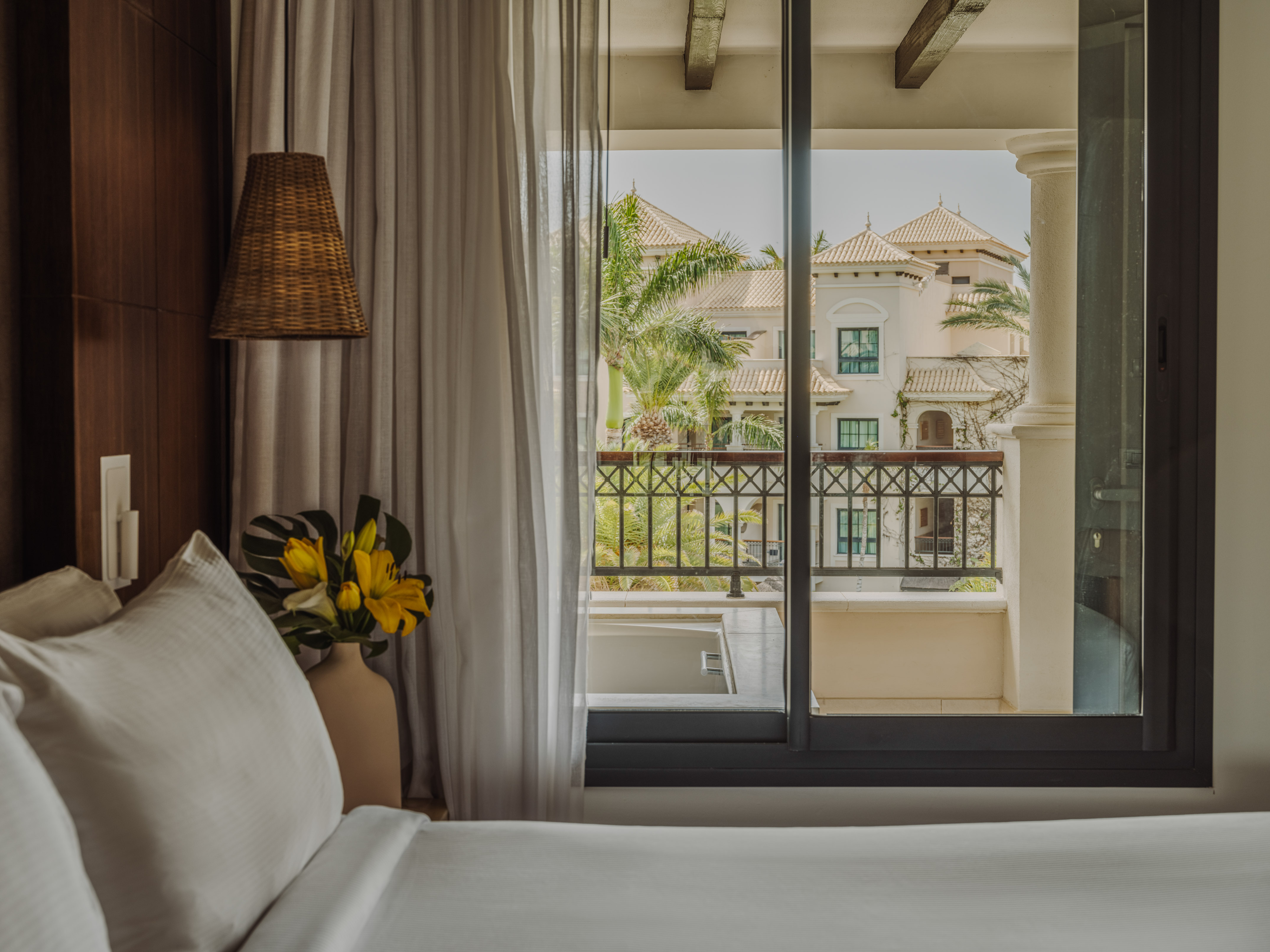 Bedroom with balcony view of white Mediterranean resort villas and palm trees.