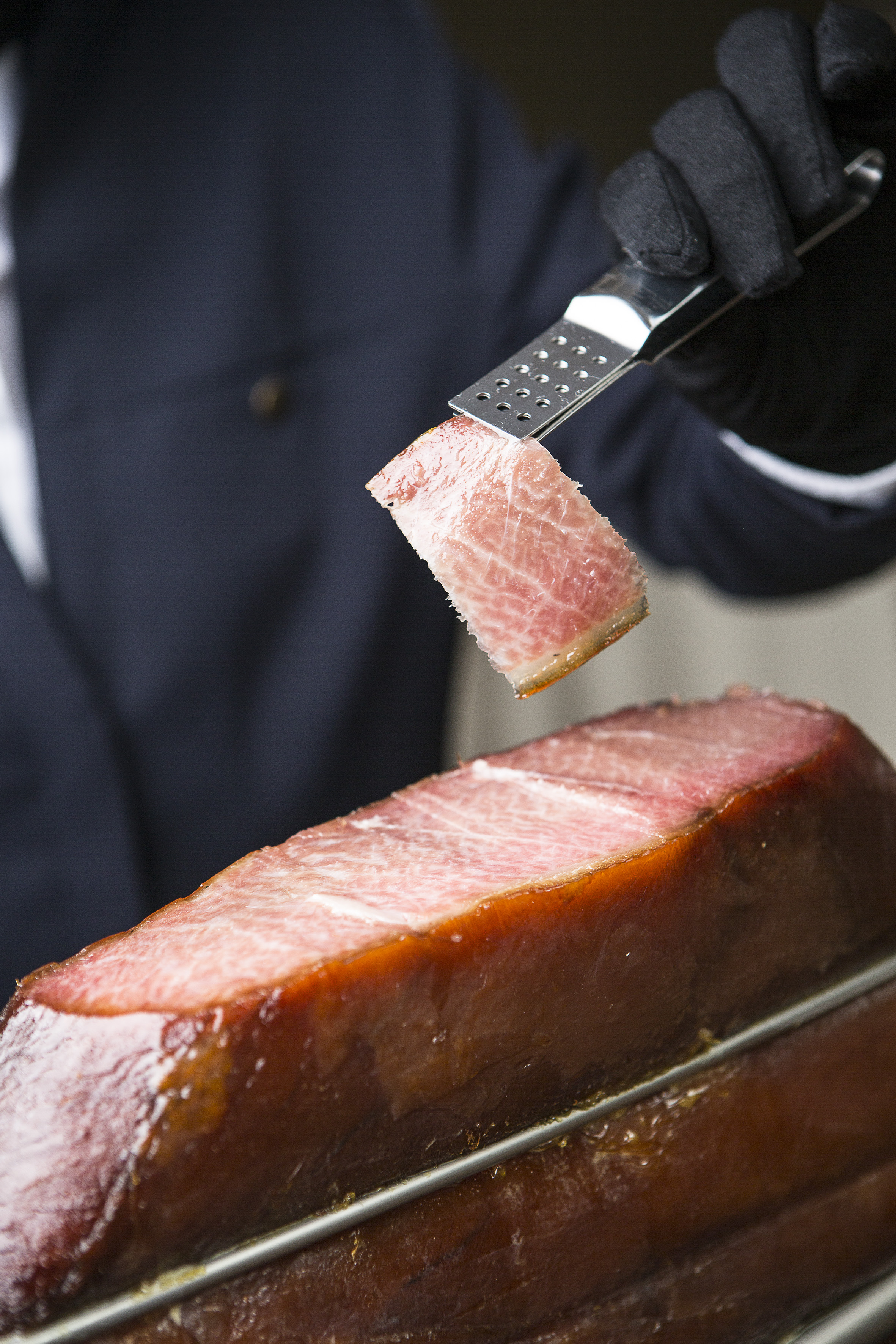Professional hand in black glove serving thin slice of cured ham with tongs.
