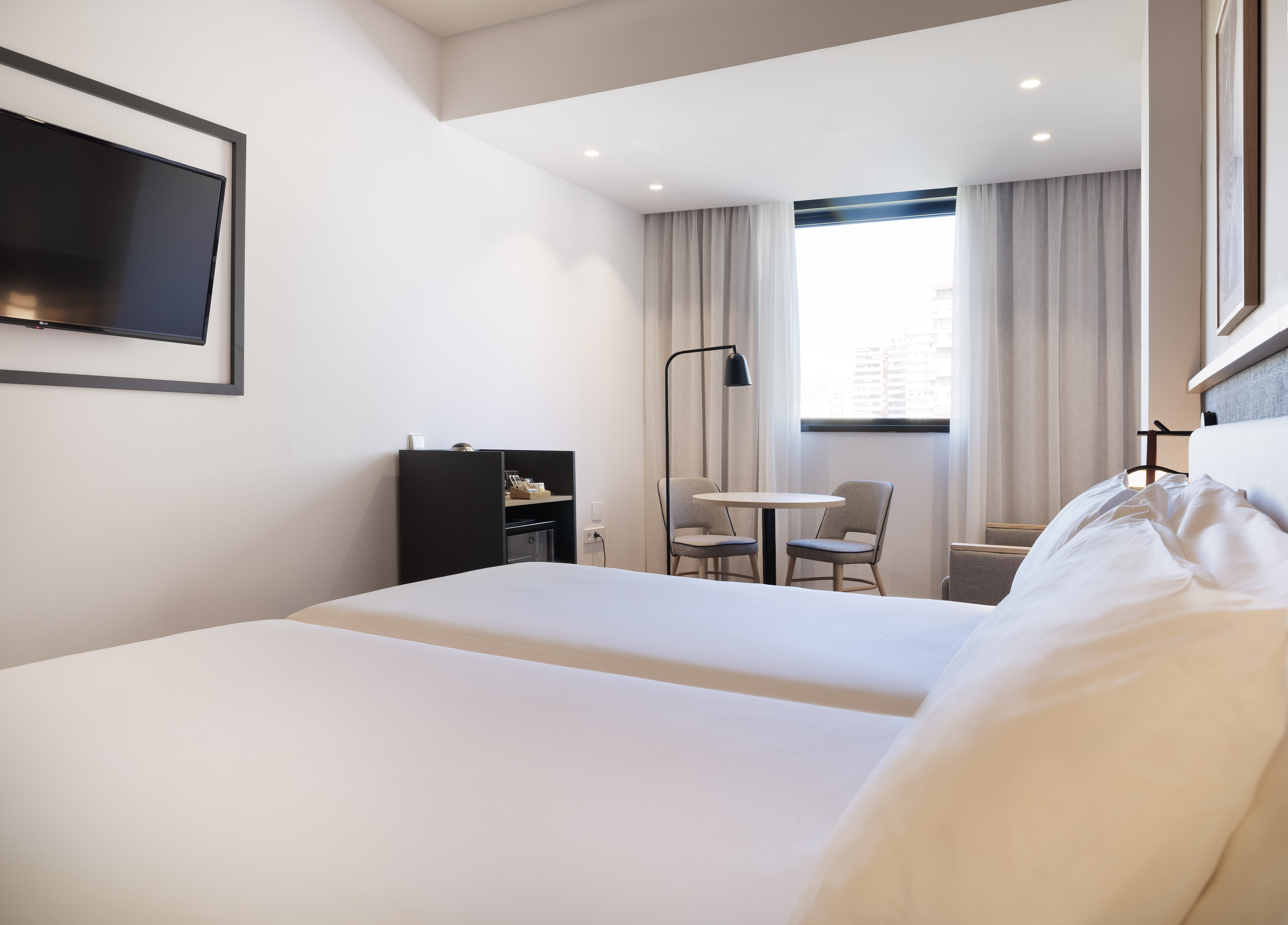 Modern hotel guest room with twin beds, wall-mounted TV, and bright window.