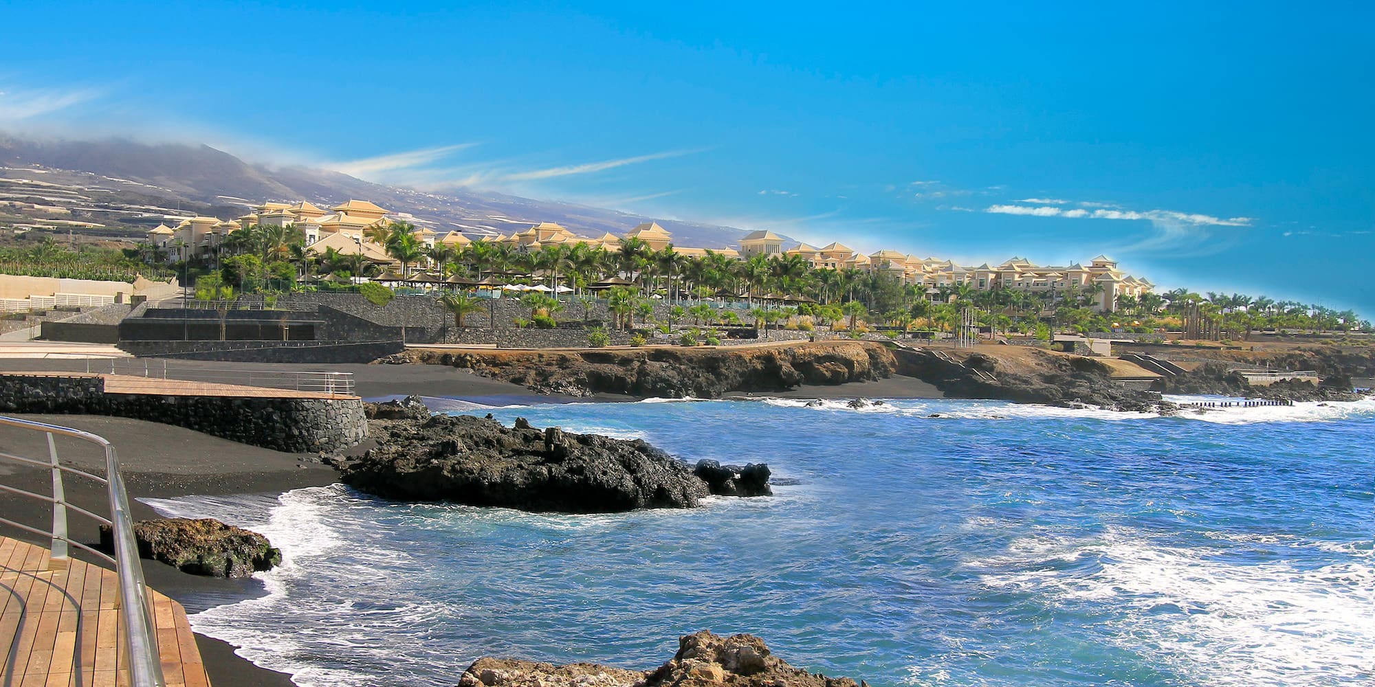 Tenerife black sand beach resort with blue ocean, palm trees, and mountains.
