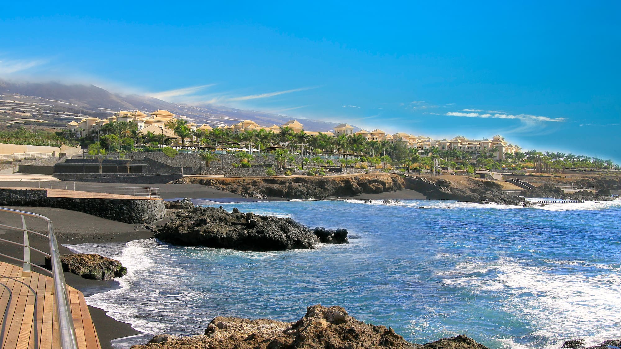 Tenerife black sand beach resort with blue ocean, palm trees, and mountains.