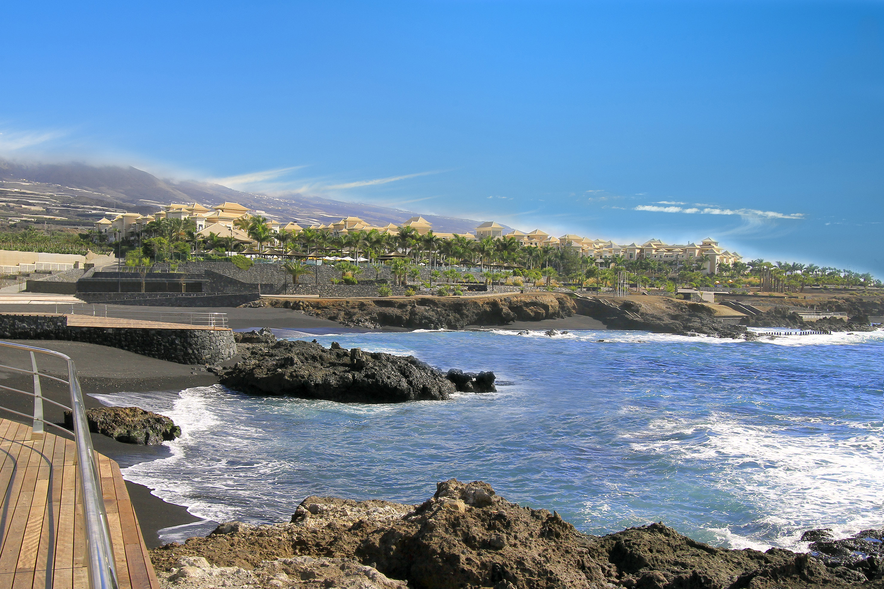Tenerife black sand beach resort with blue ocean, palm trees, and mountains.