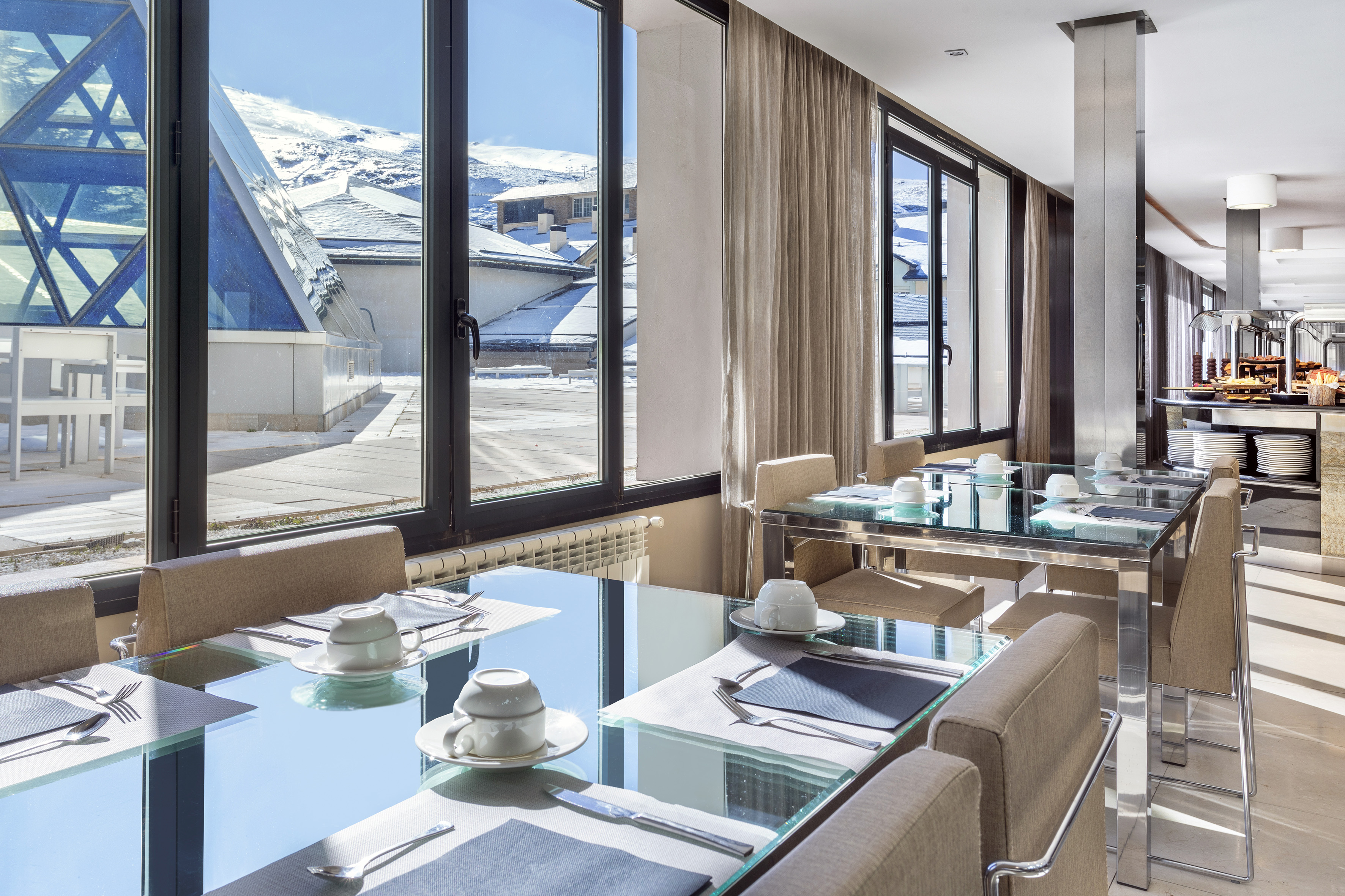 Modern dining room with snowy mountain view, glass tables, and breakfast buffet.