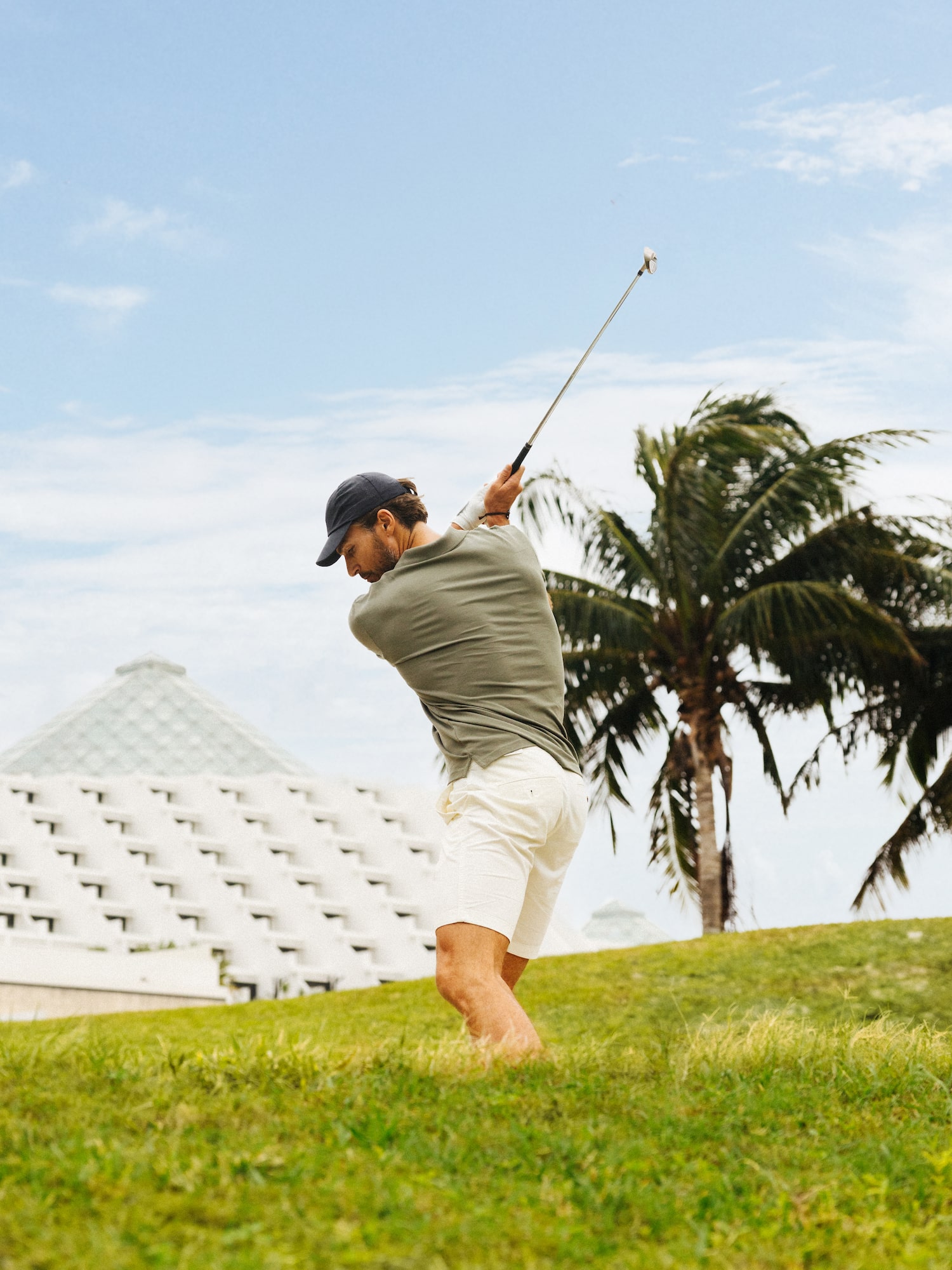 Golfista haciendo swing en campo tropical con palmeras y hotel.