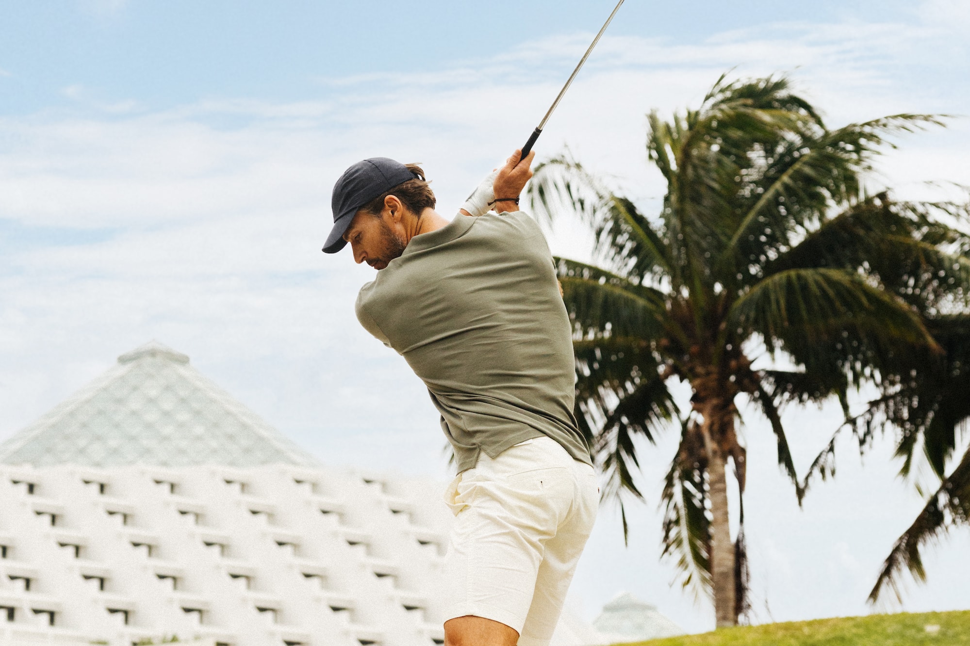 Golfer in olive polo and white shorts swings on a sunny course.