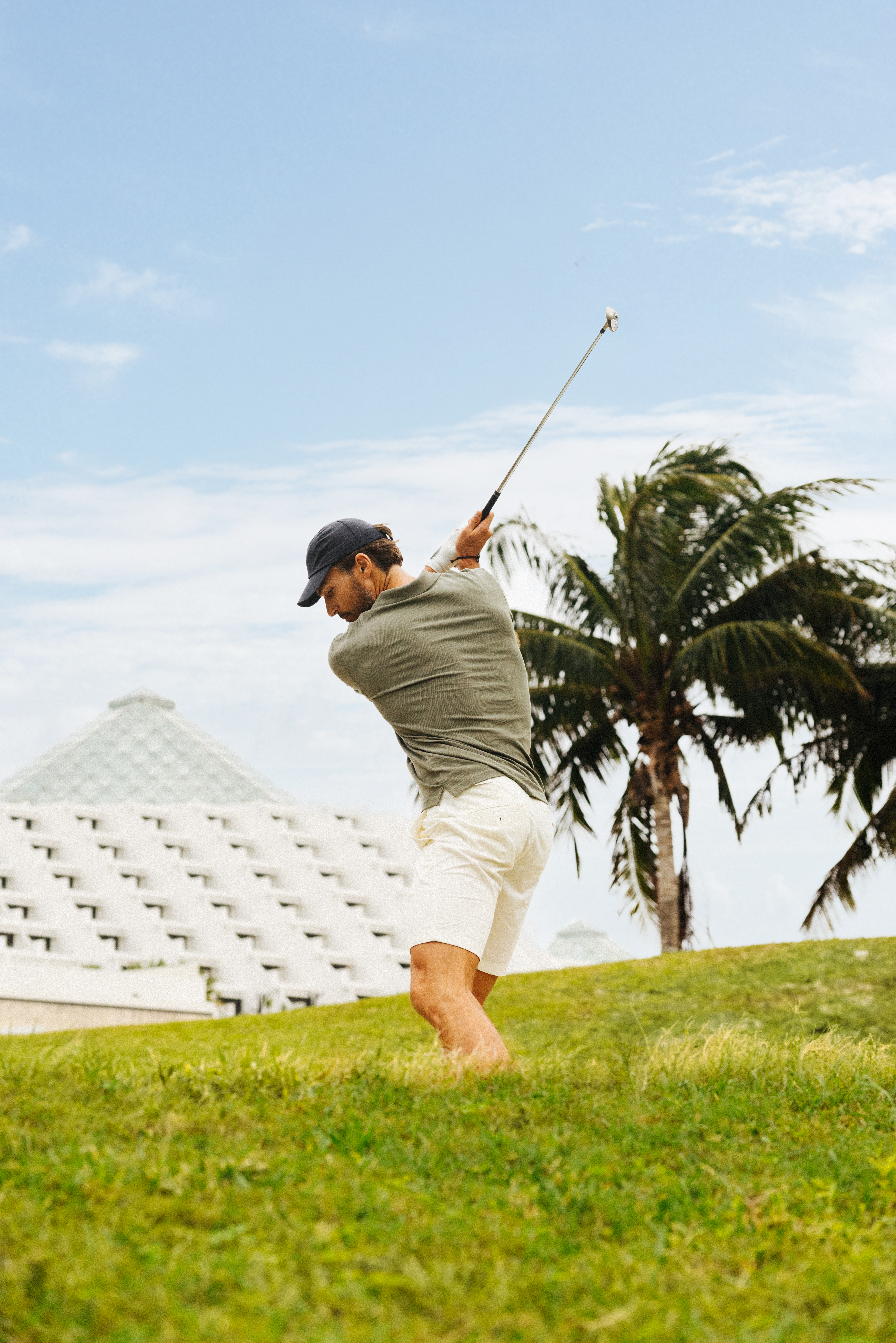 Golfer in olive polo and white shorts swings on a sunny course.
