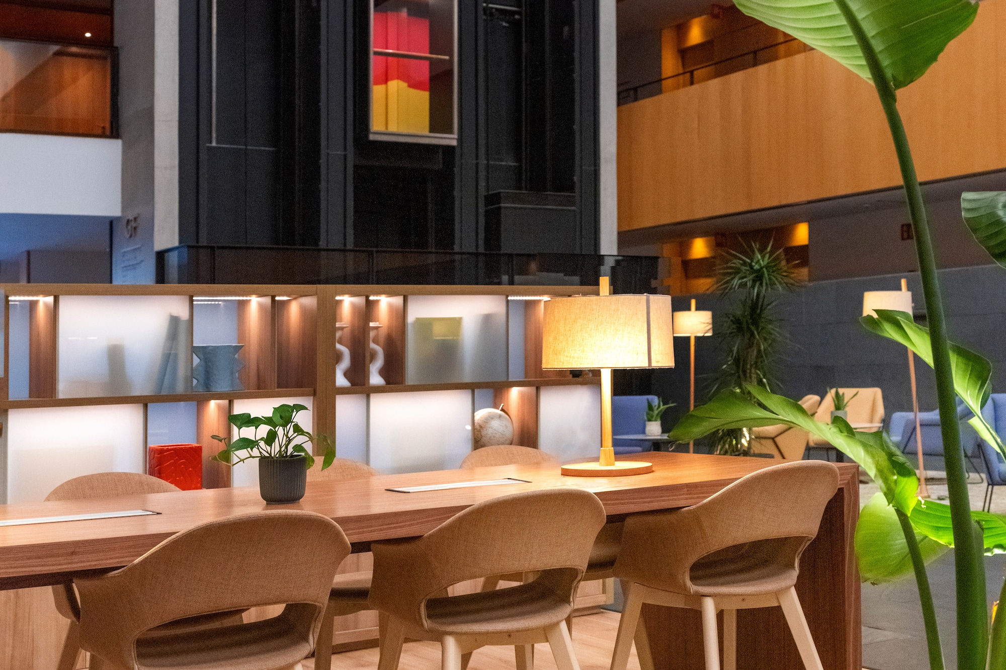 Modern hotel lobby workspace with communal table, chairs, and atrium elevators.