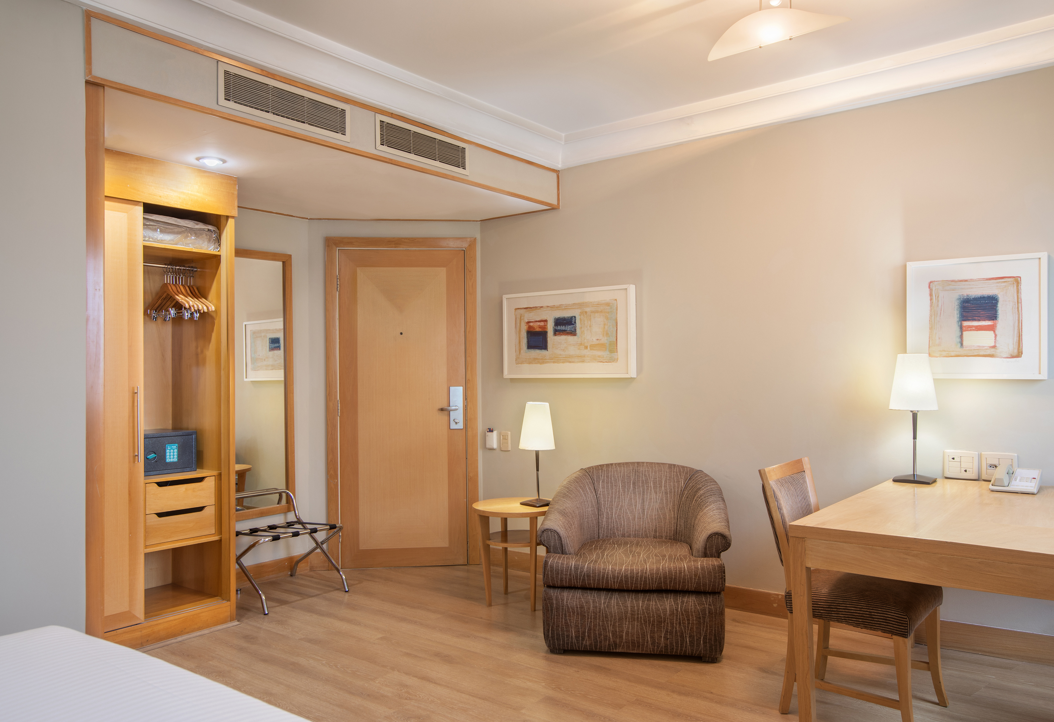 Bright hotel room featuring a wardrobe, safe, desk, and armchair.