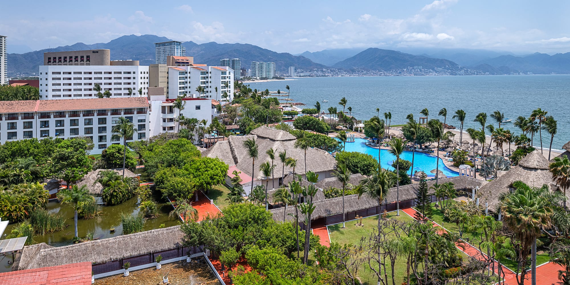 Aerial view of a beachfront resort with pools, palm trees, ocean, and mountains.