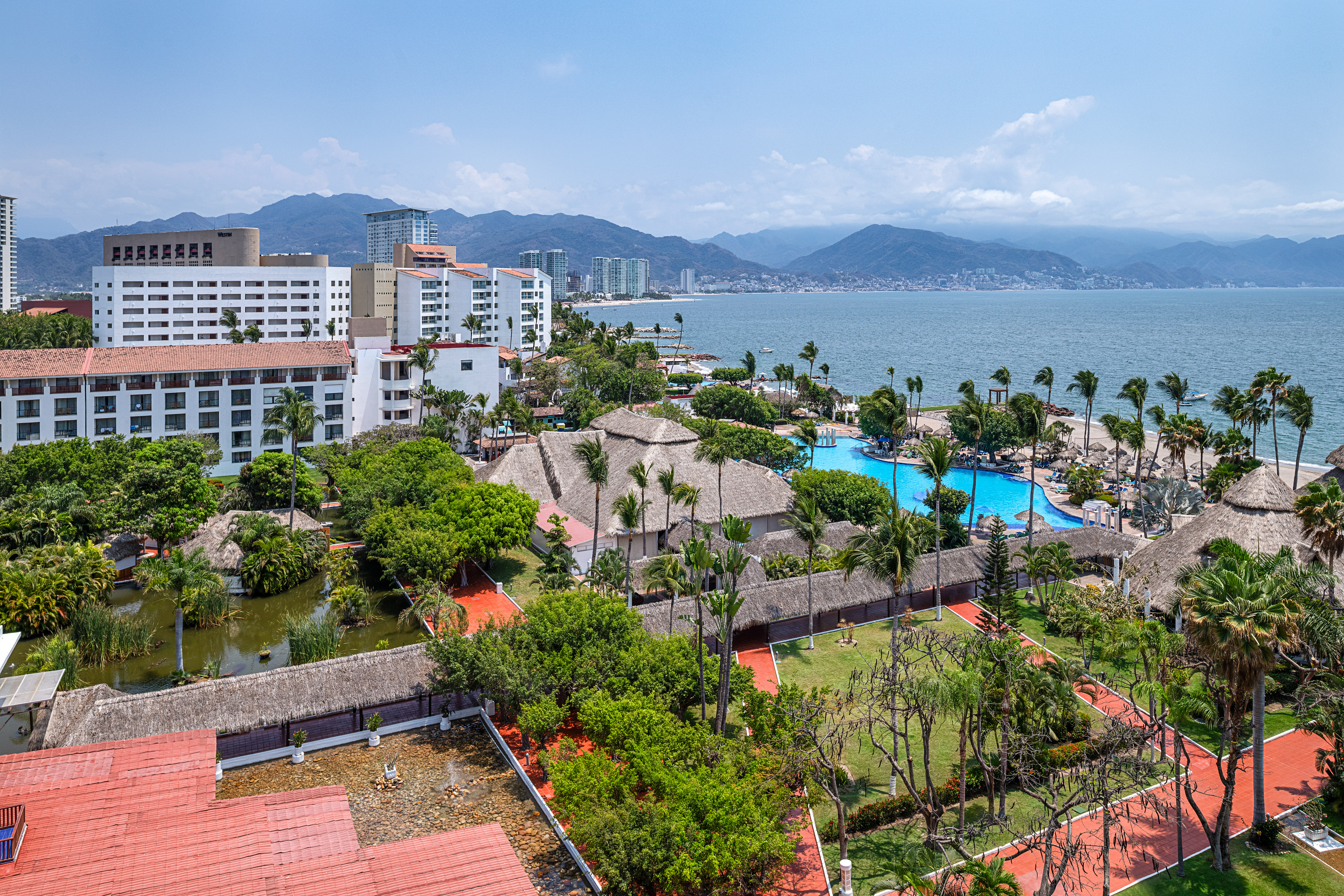 Aerial view of a beachfront resort with pools, palm trees, ocean, and mountains.