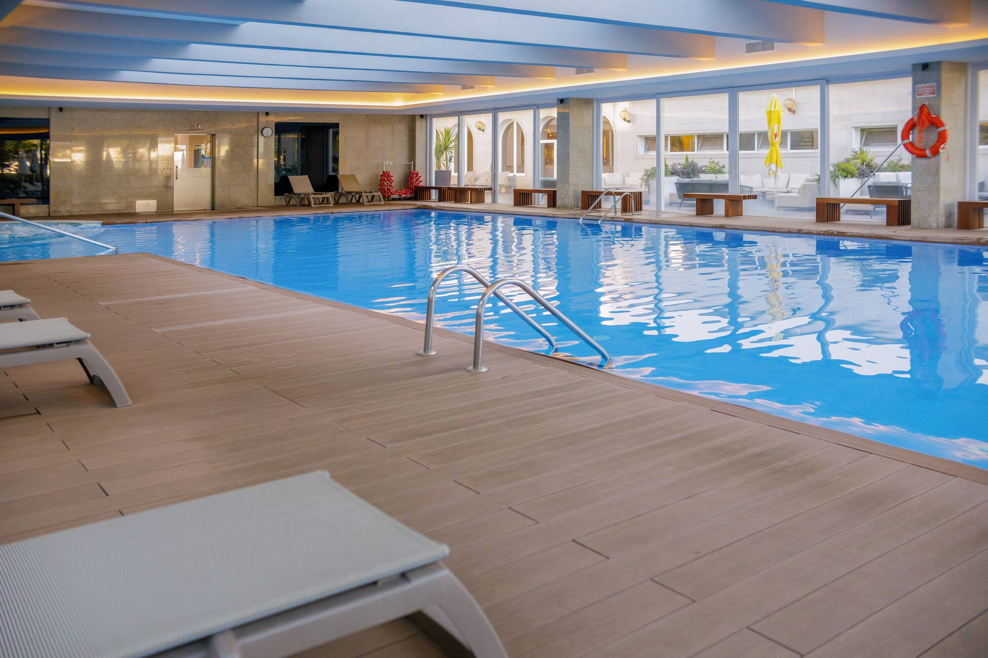 Modern indoor hotel pool with blue water, wooden deck, and sun loungers.