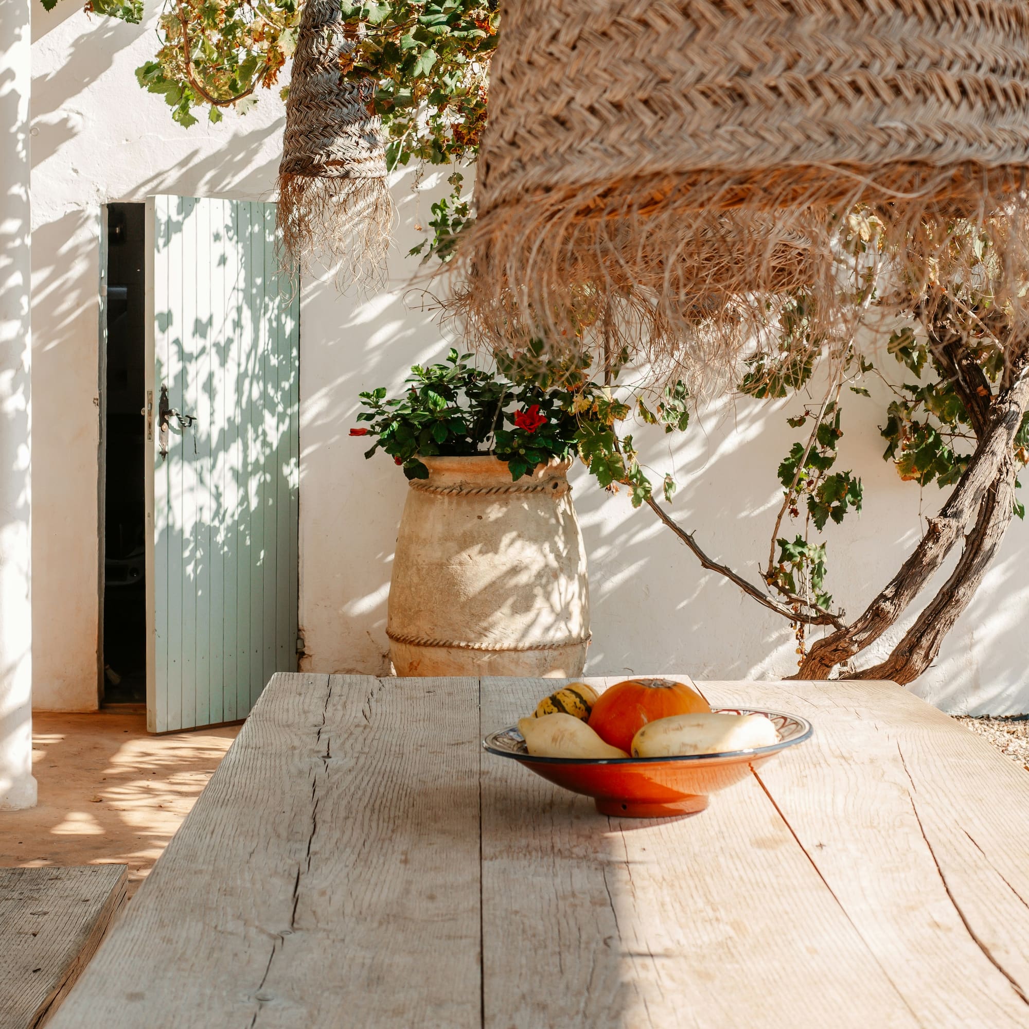 Sunlit Mediterranean patio with rustic wooden table, gourds, and a woven straw lamp.