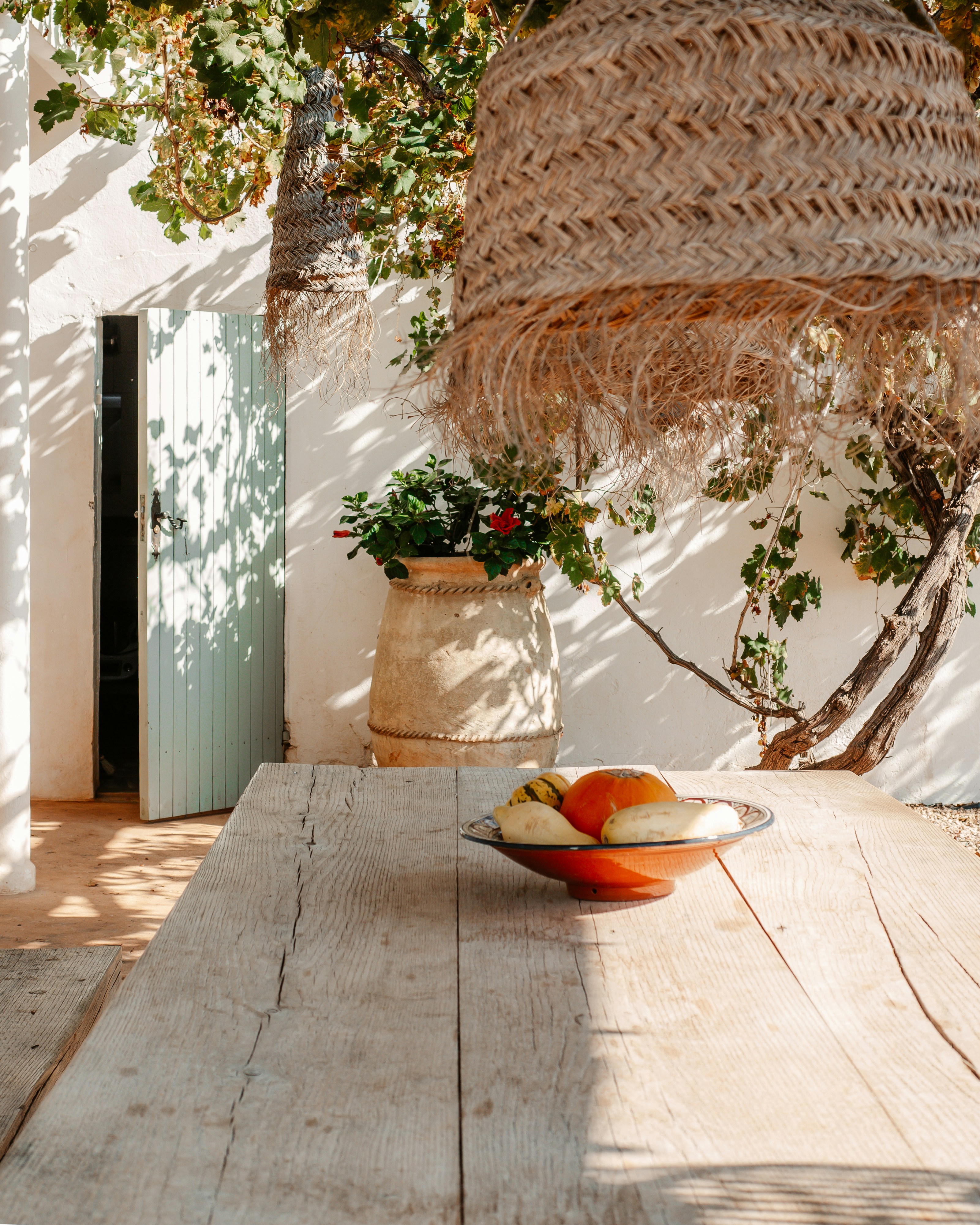 Sunlit Mediterranean patio with rustic wooden table, gourds, and a woven straw lamp.