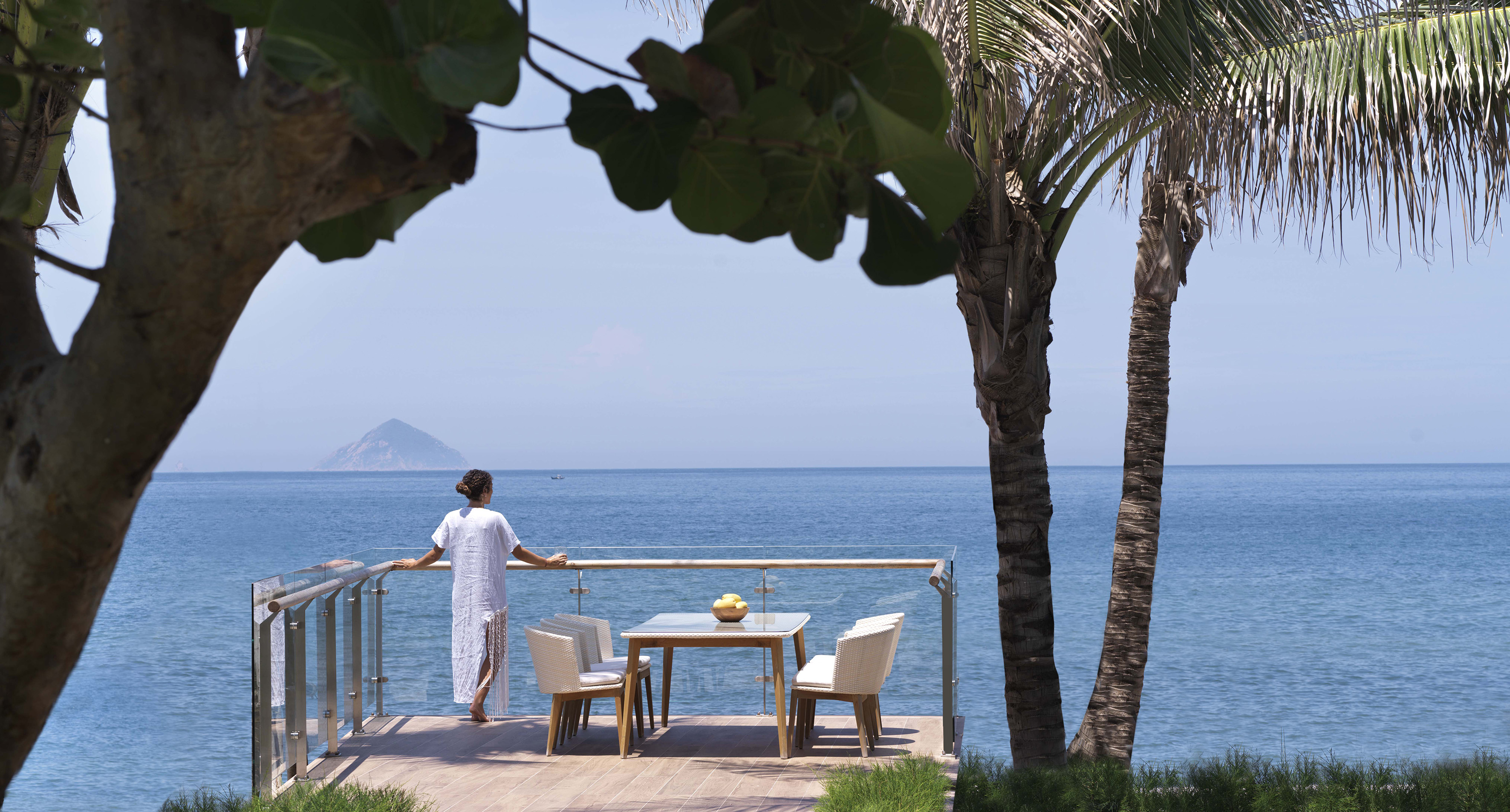 Woman on tropical balcony enjoying ocean view with dining set and distant island.