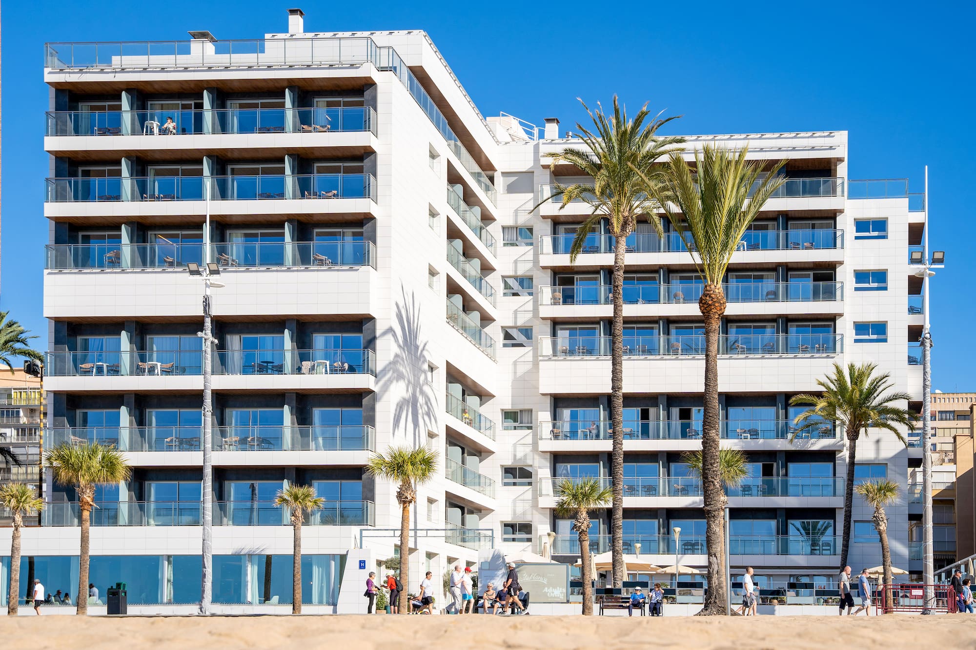 Modern white beachfront hotel with balconies and palm trees.