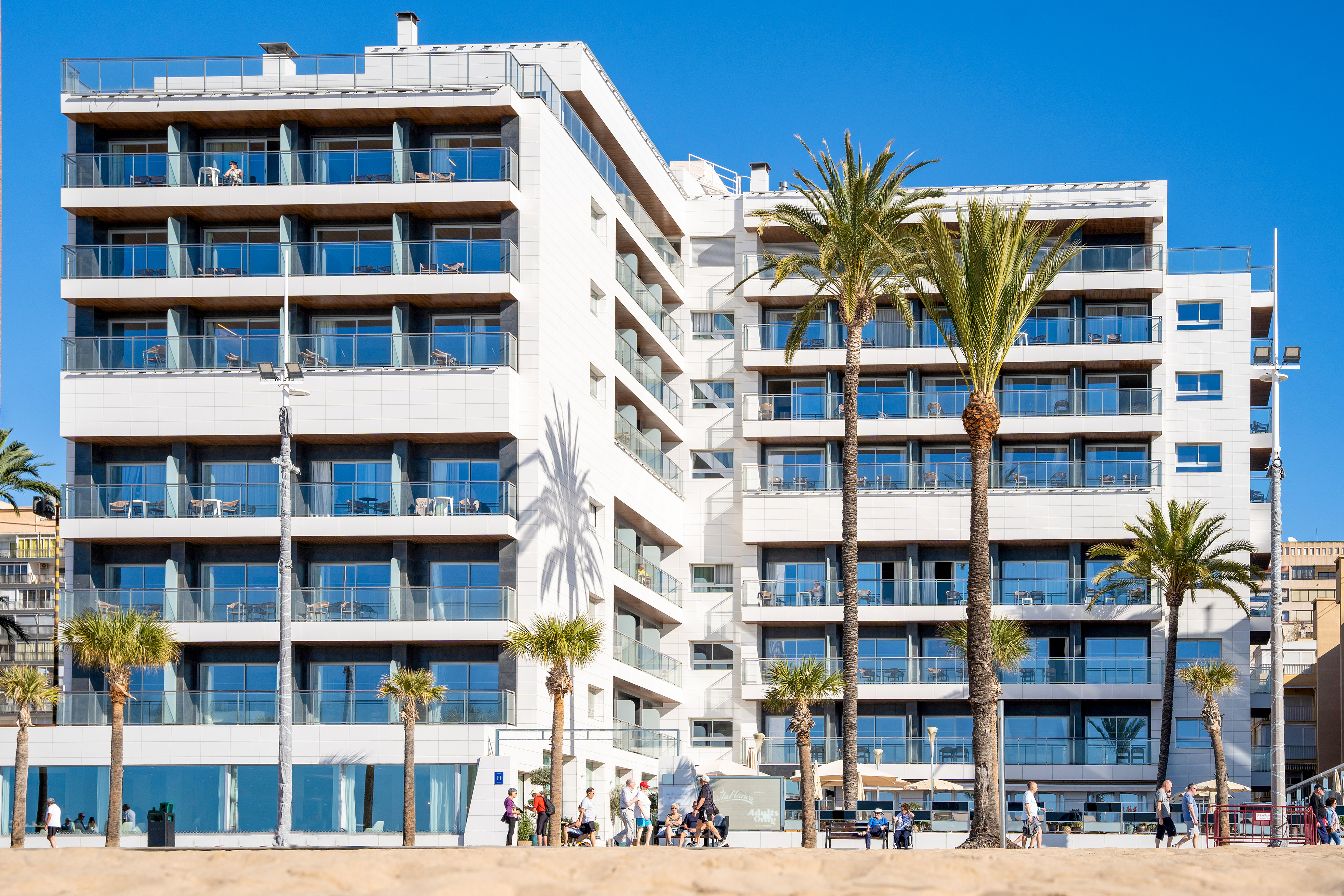 Modern white beachfront hotel with balconies and palm trees.