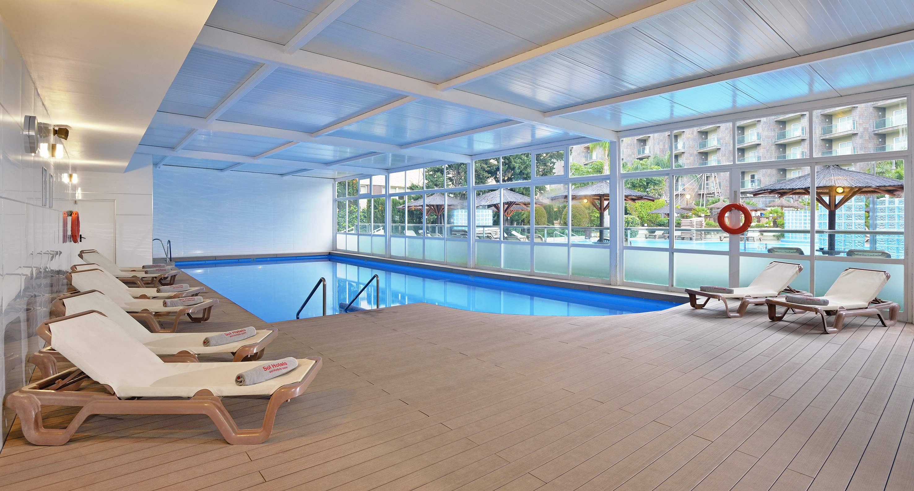 Indoor hotel pool with lounge chairs, wooden deck, and large windows to outdoor resort.