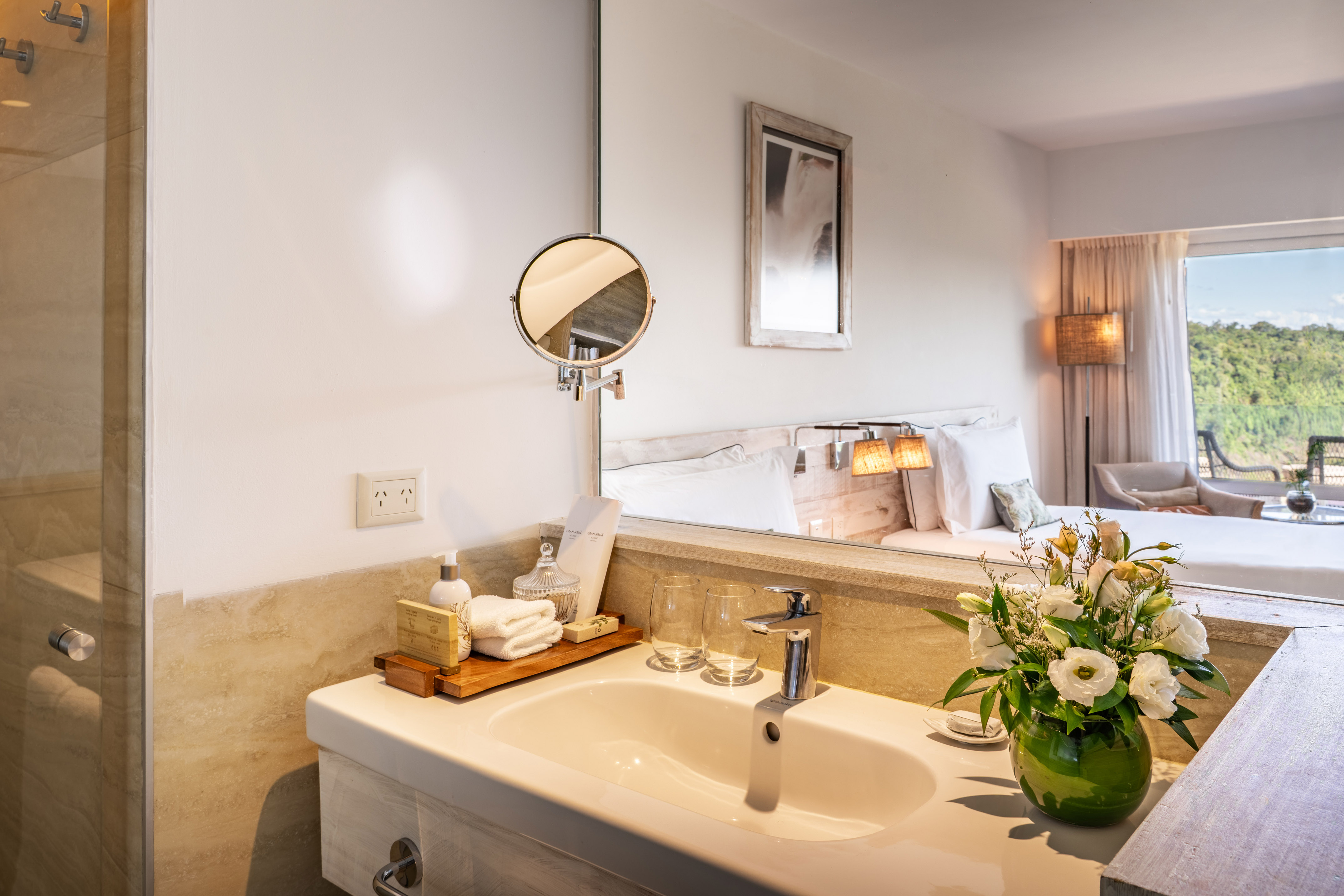 Modern hotel bathroom vanity with sink, mirror, toiletries, and reflected bedroom view.