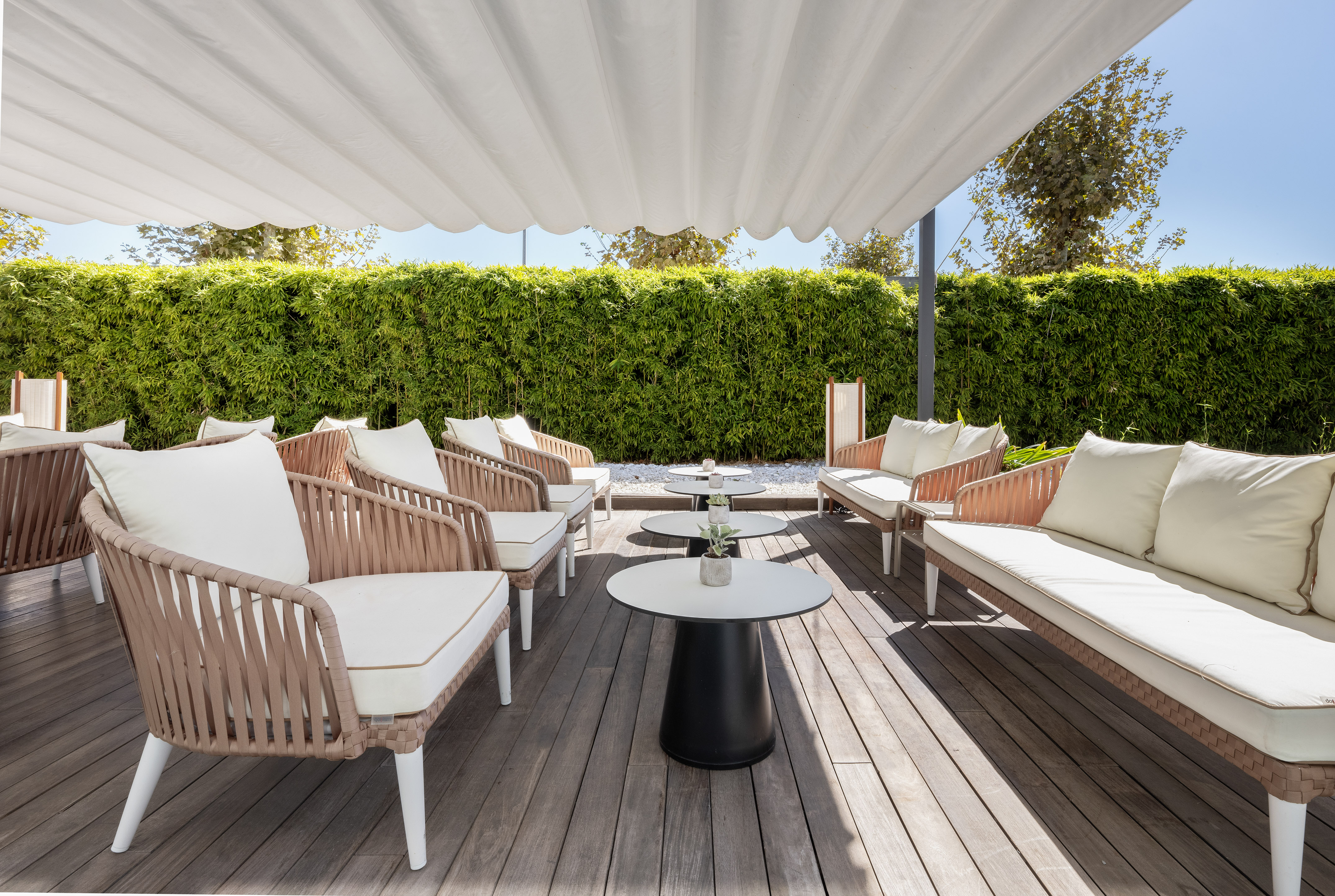 Sunny outdoor terrace with white canopy, rattan seating, wooden deck, and green wall.