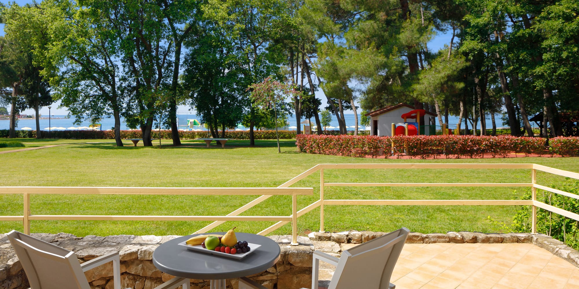 a table with fruit on it and chairs on a patio with a lawn and trees