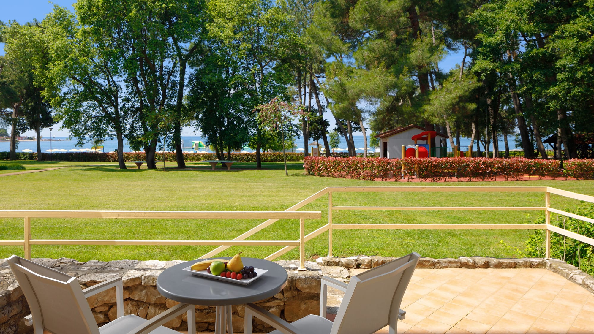 a table with fruit on it and chairs on a patio with a lawn and trees