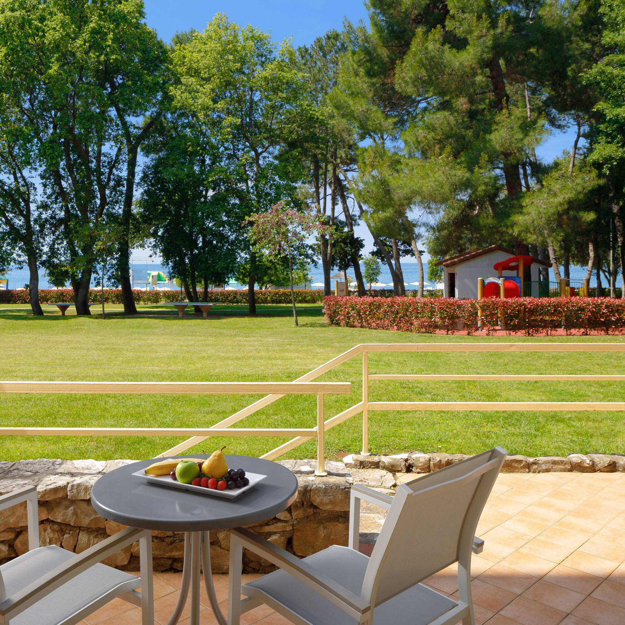 a table with fruit on it and chairs on a patio with a lawn and trees