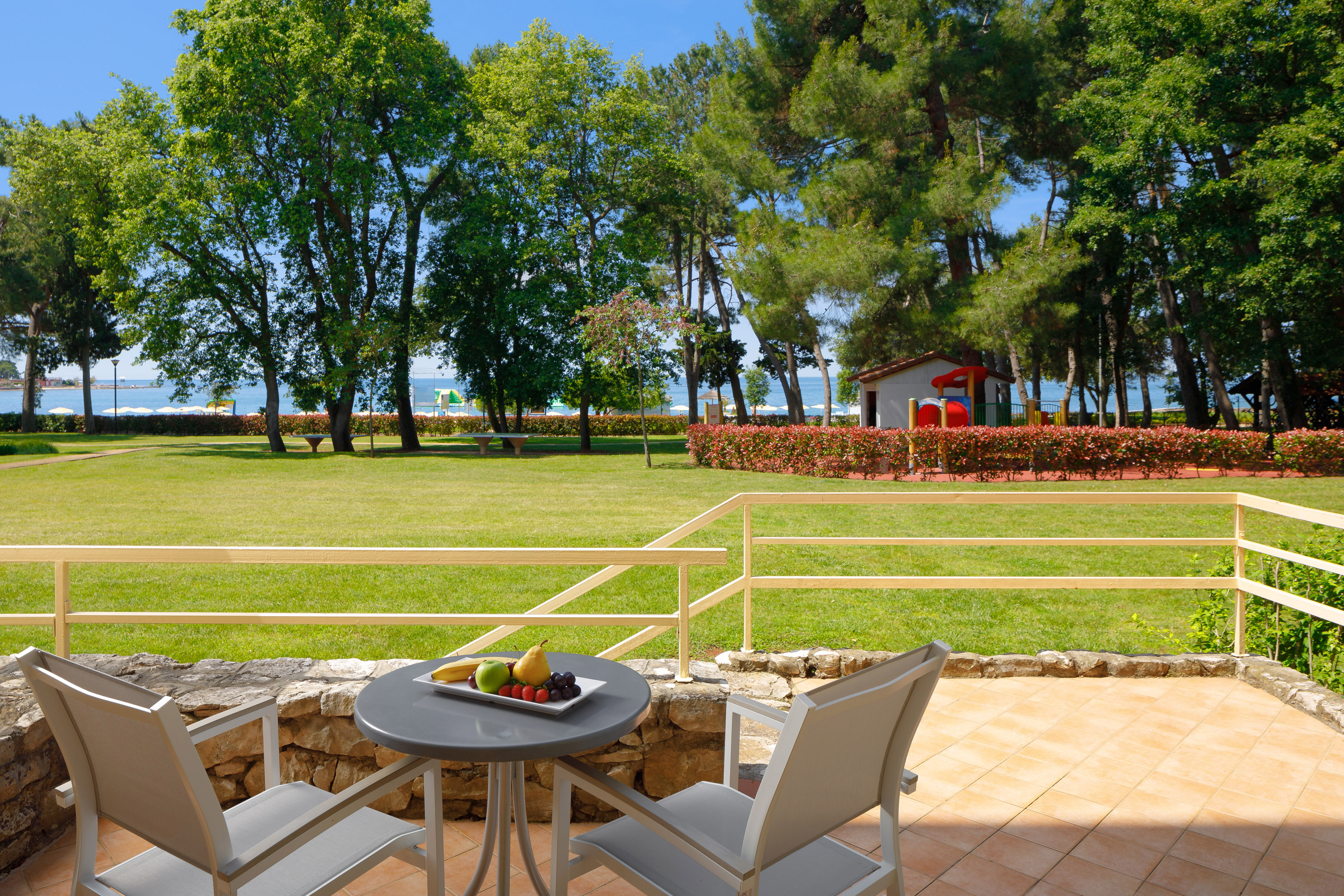 a table with fruit on it and chairs on a patio with a lawn and trees