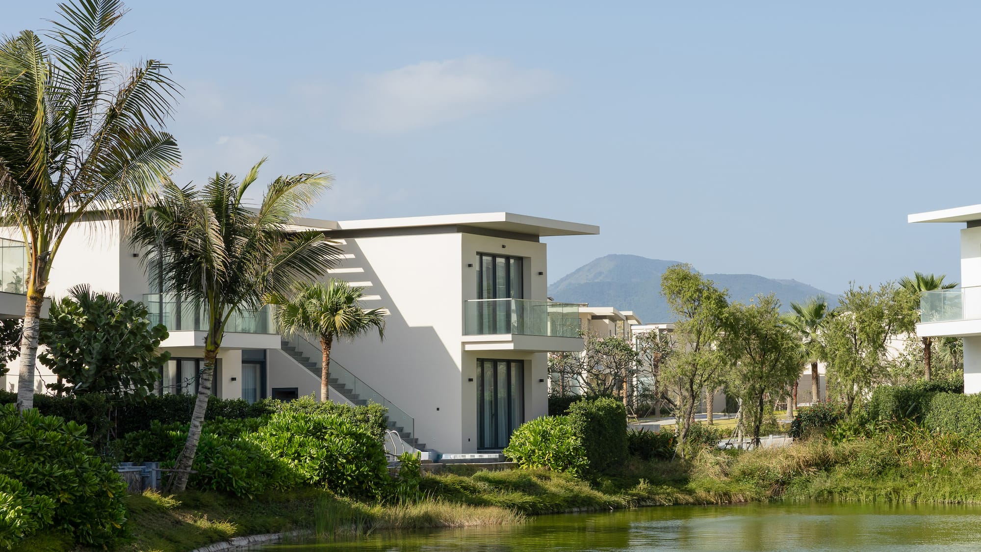 Modern resort villa by tranquil lake, lush palm trees, mountain view.