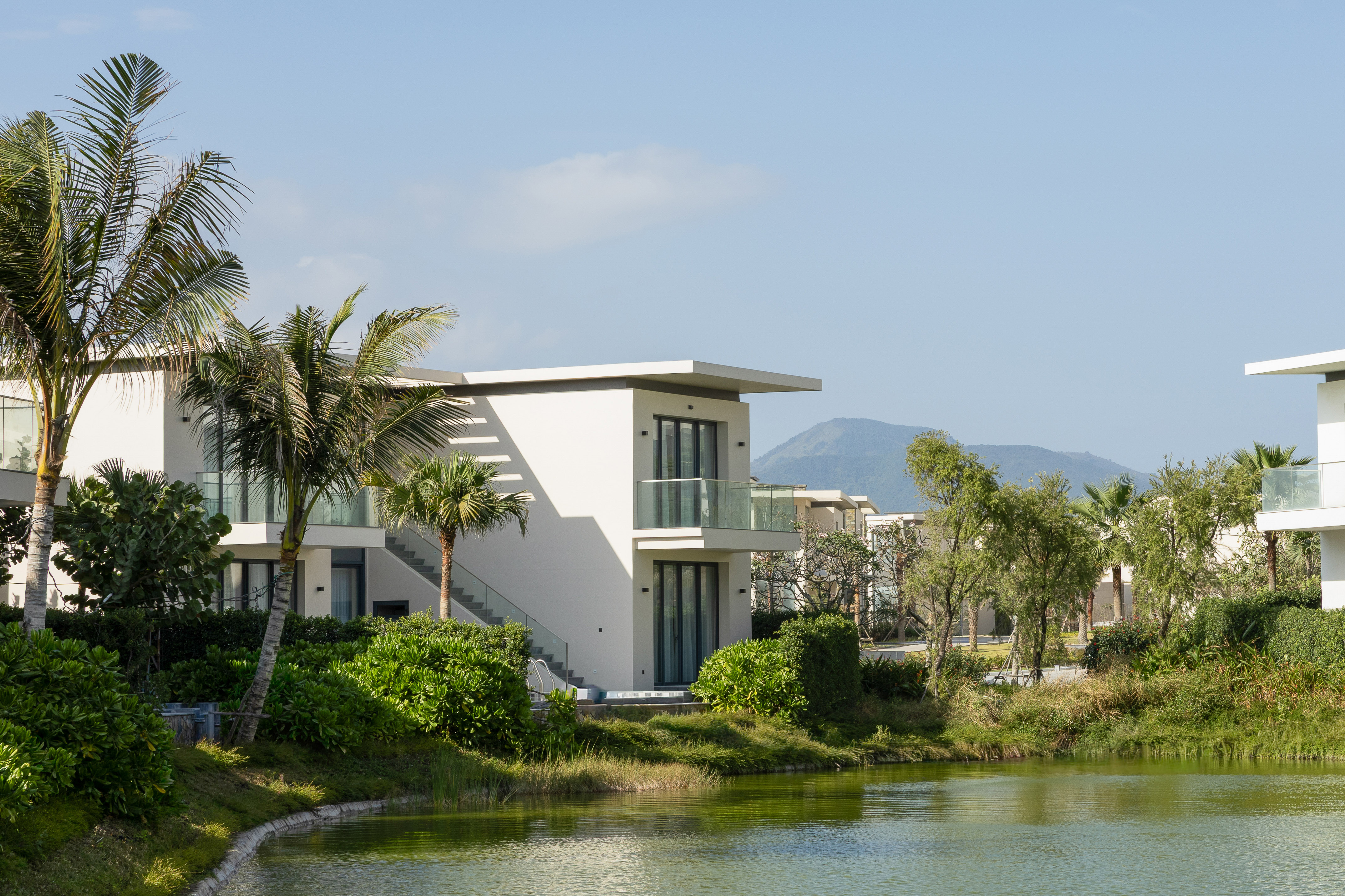 Modern resort villa by tranquil lake, lush palm trees, mountain view.