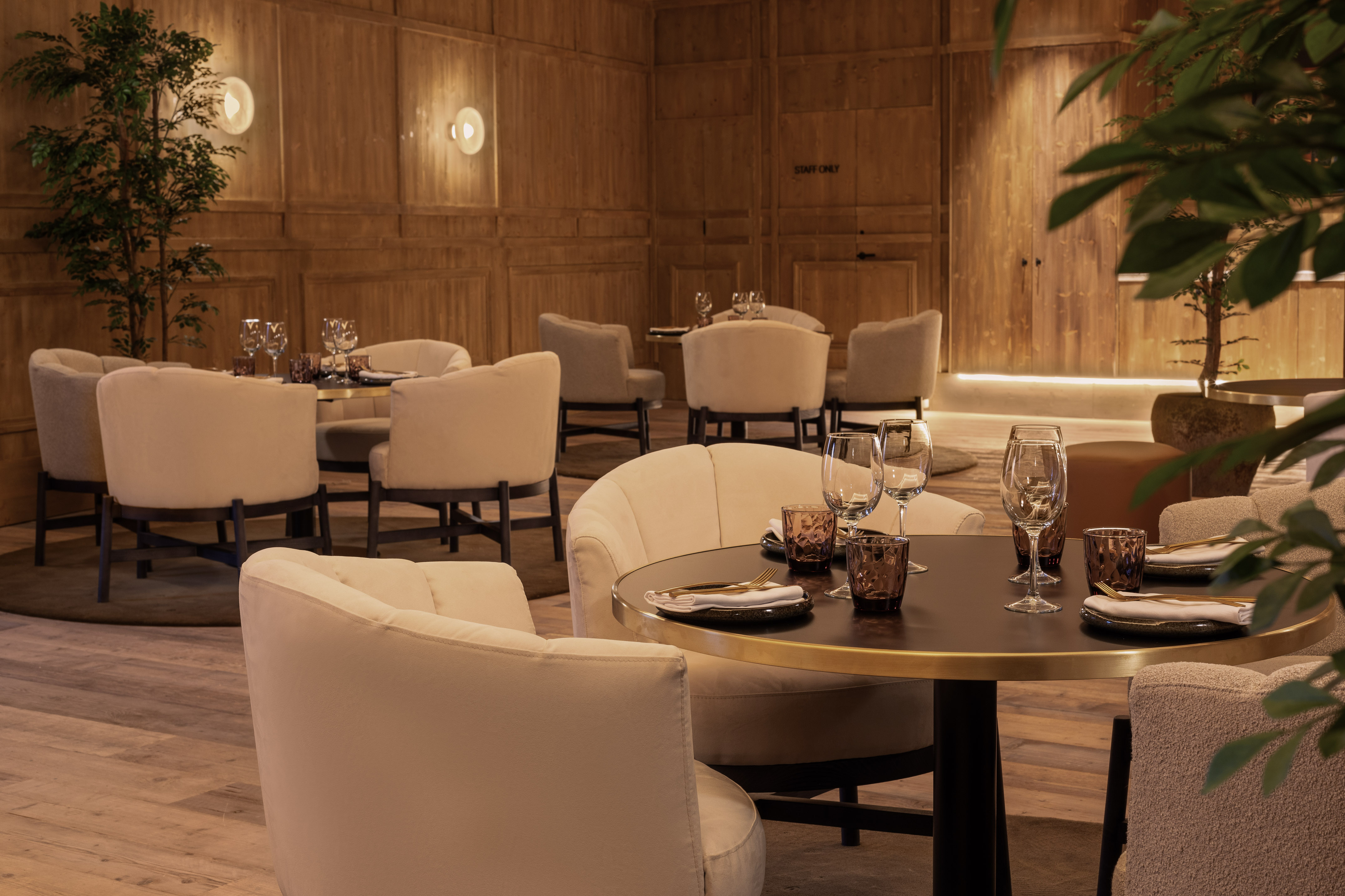 Modern restaurant interior with warm wood panels, elegant cream chairs, and set tables.