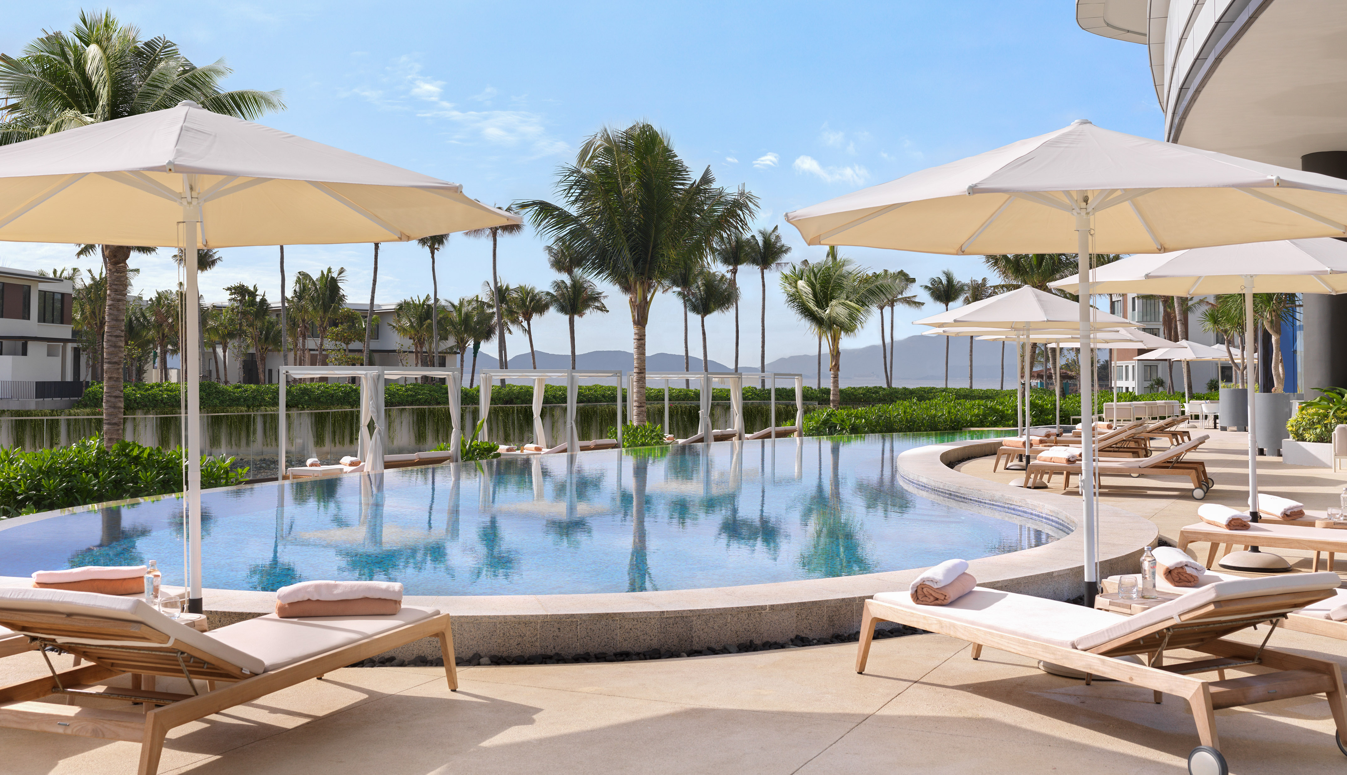 Outdoor resort pool with sun loungers, umbrellas, and palm trees.