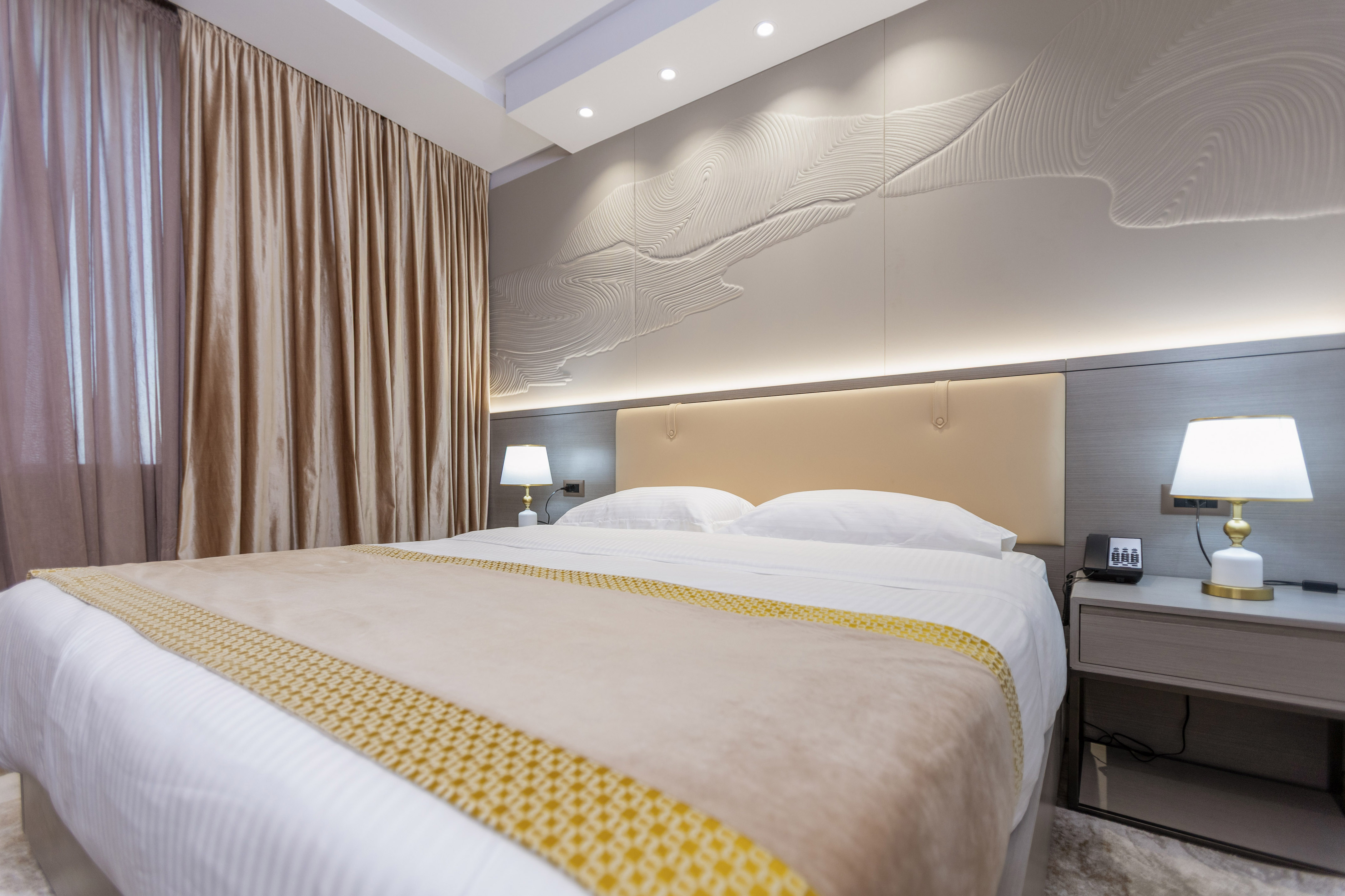 Modern hotel bedroom with king bed, beige linens, and textured wall art.