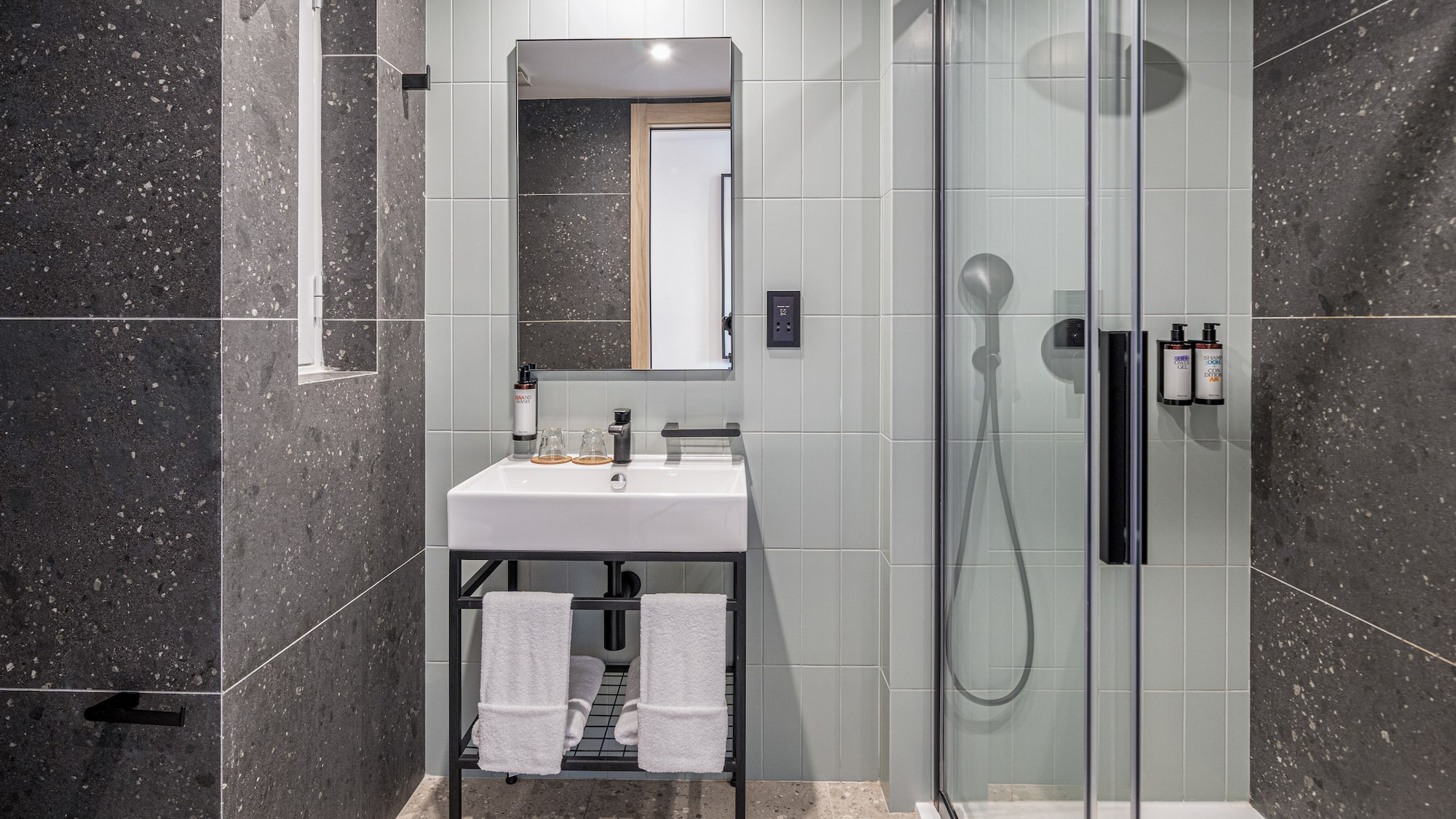 Modern hotel bathroom with vanity, mirror, walk-in shower, and dark tiled walls.