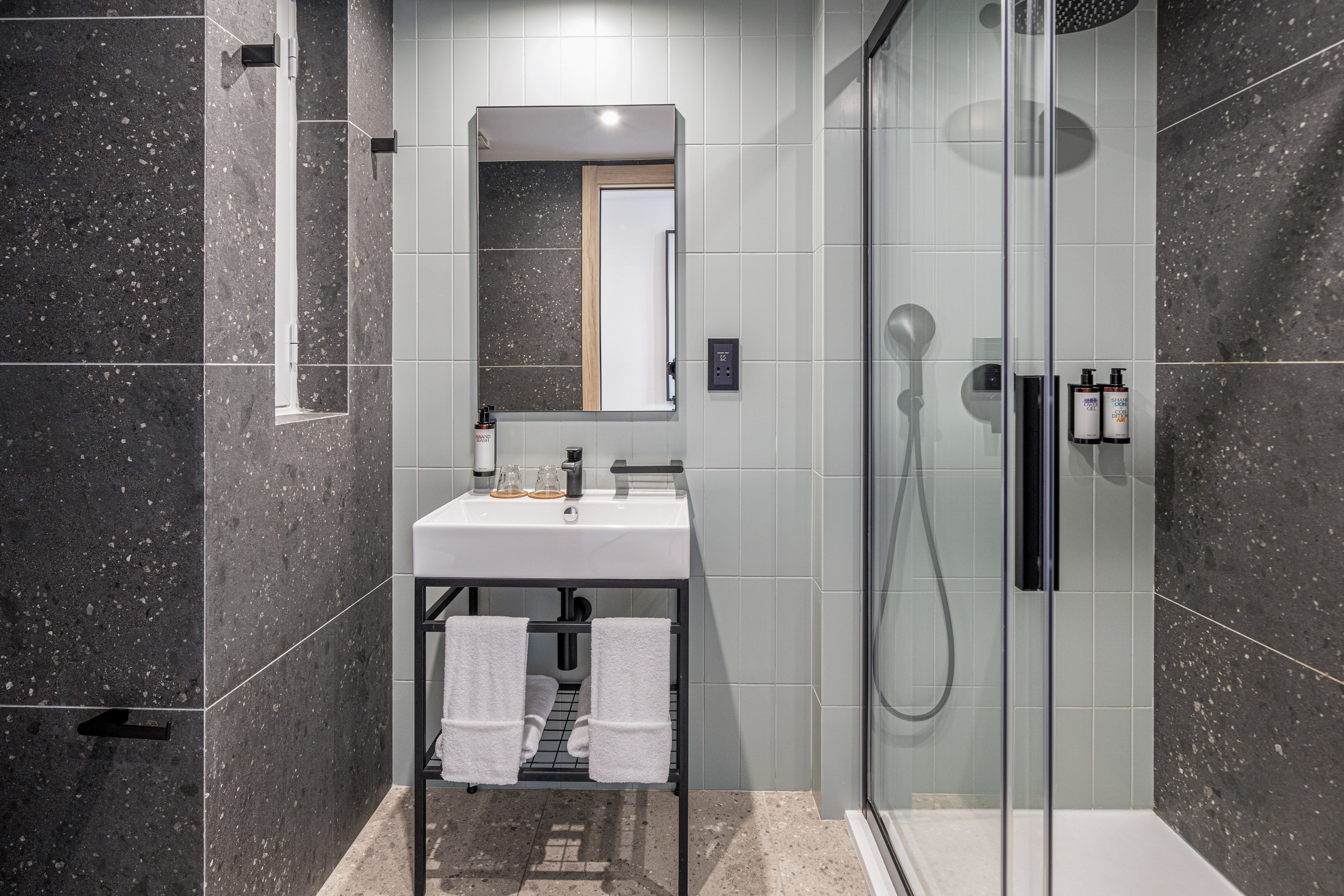 Modern hotel bathroom with vanity, mirror, walk-in shower, and dark tiled walls.