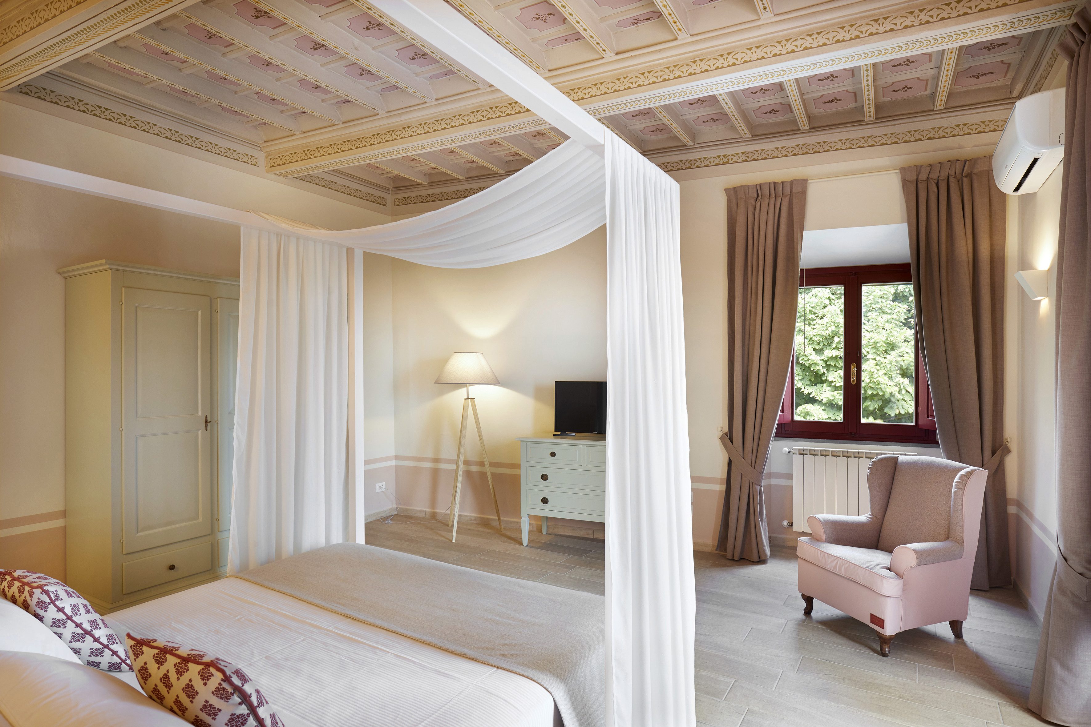 Elegant bedroom with four-poster bed, ornate painted ceiling, and pink armchair.