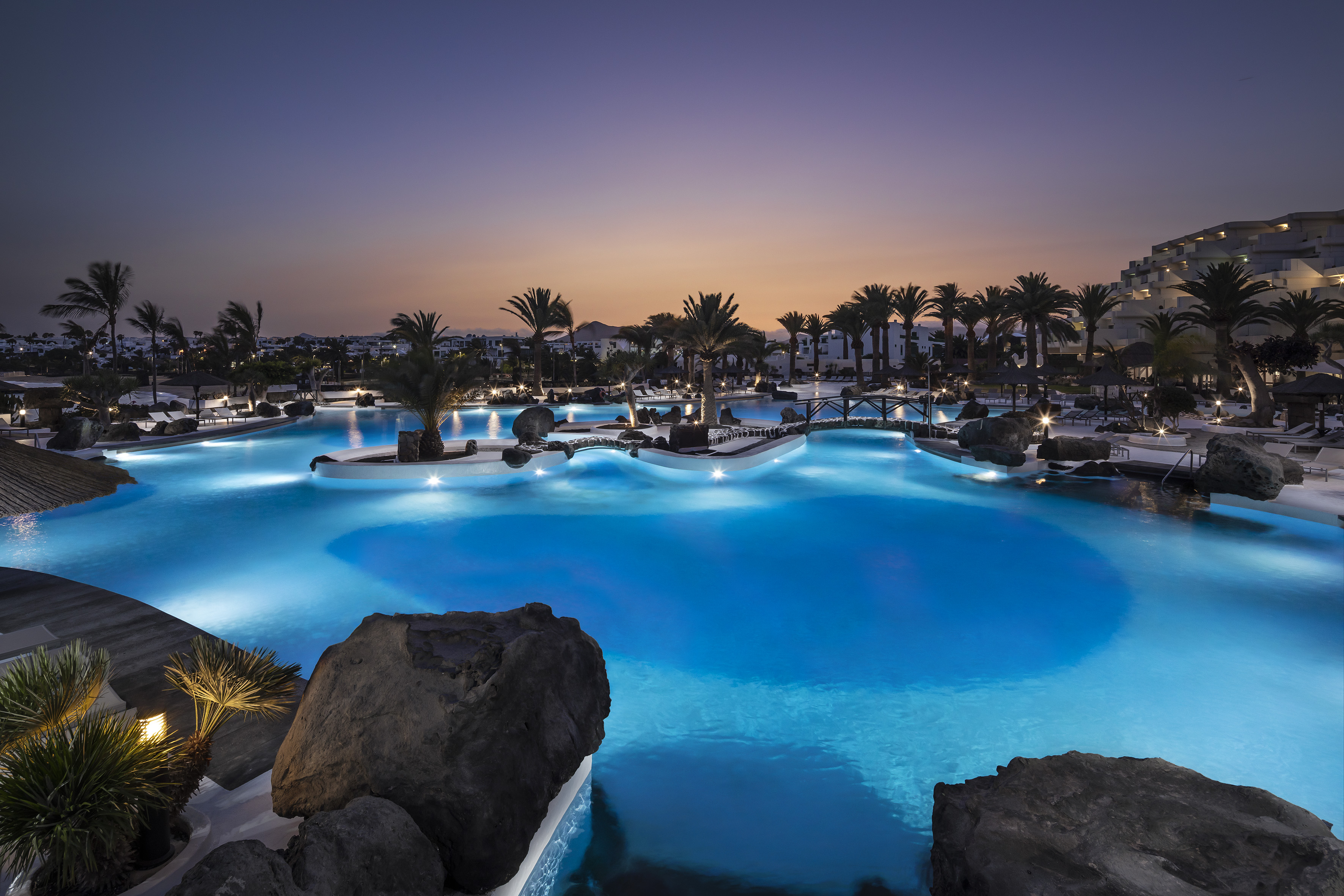 Blue illuminated resort pool, tropical palm trees, and hotel at sunset.
