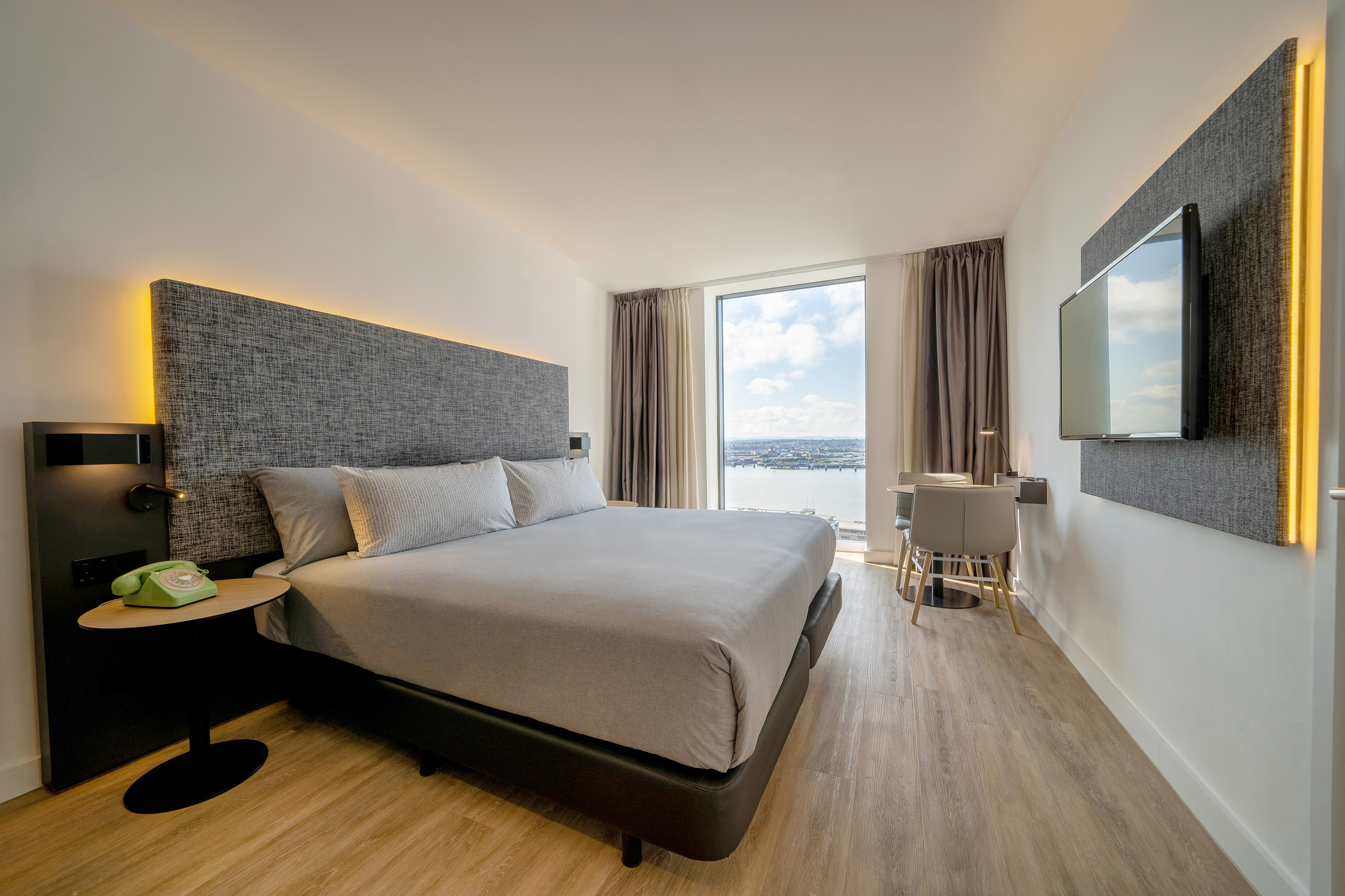 Modern hotel room with king bed, illuminated headboard, and city waterfront view.