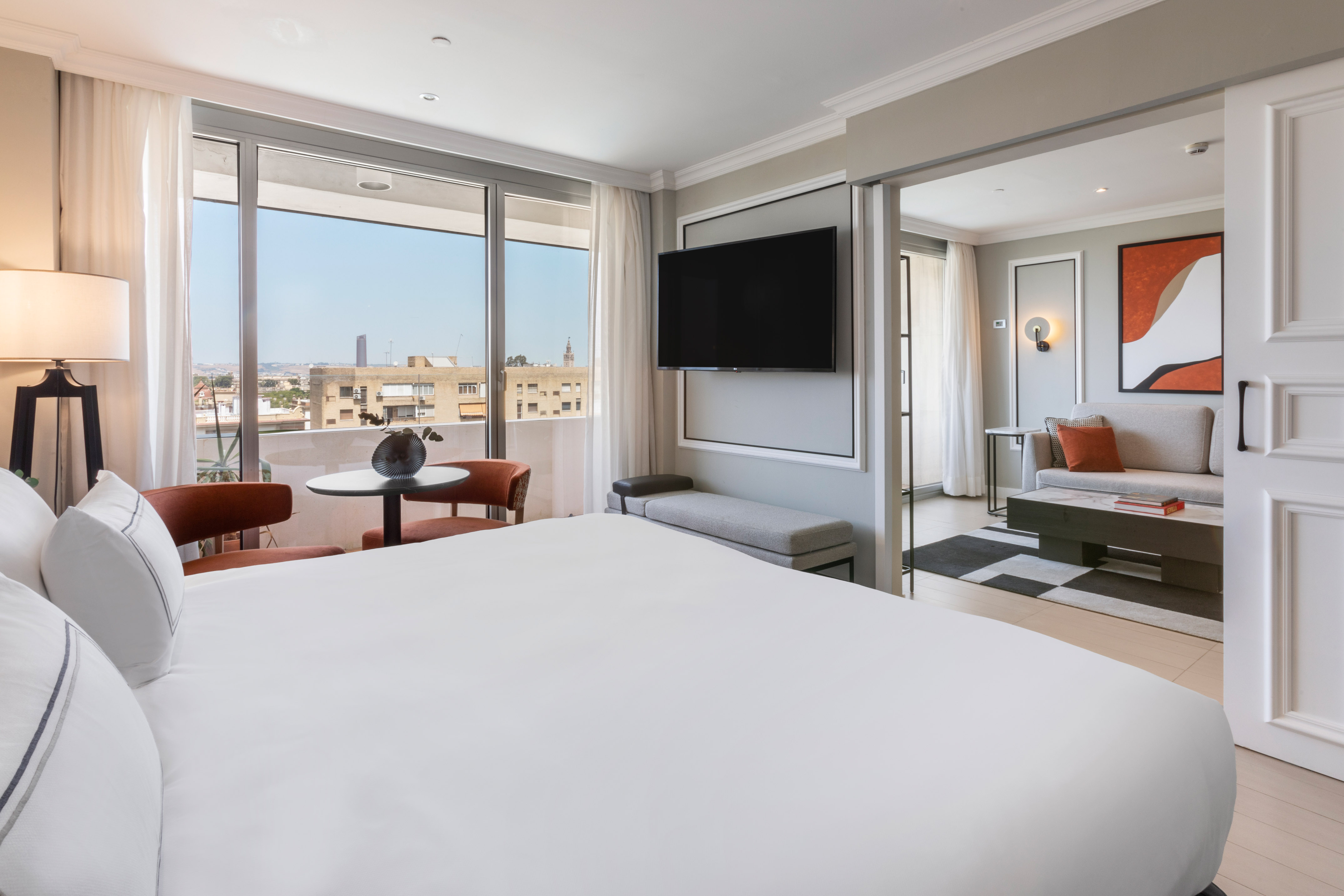 Modern hotel suite with bedroom, city view balcony, and separate living area.