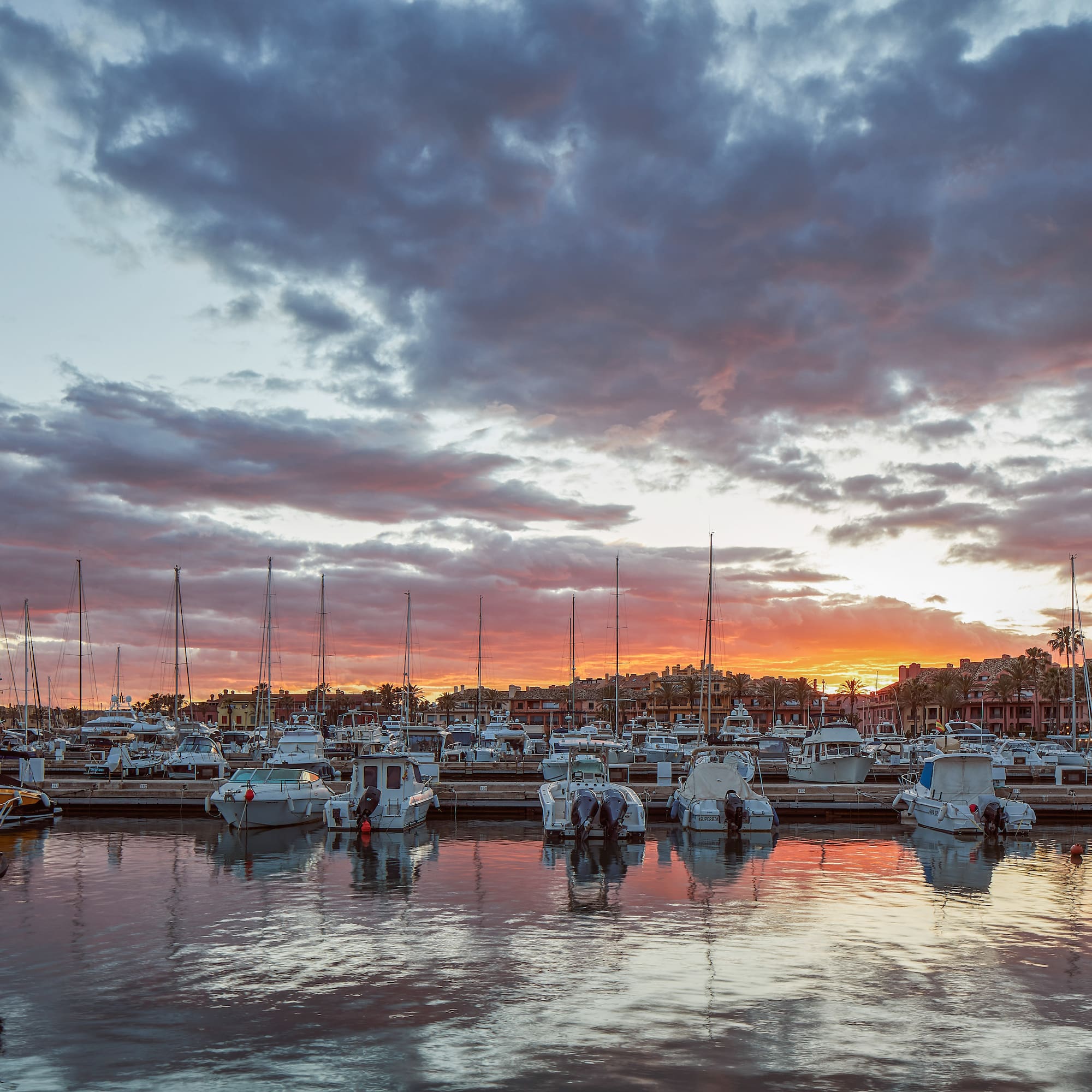 Marina full of boats at vibrant sunset with cloudy sky and water reflections.