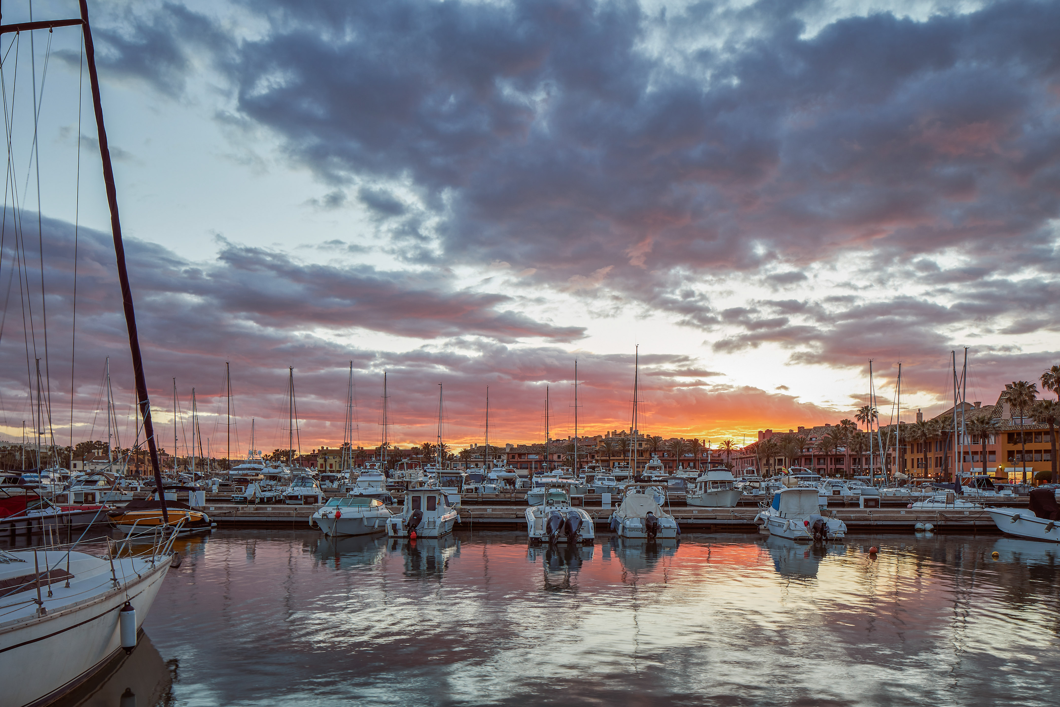 Marina full of boats at vibrant sunset with cloudy sky and water reflections.